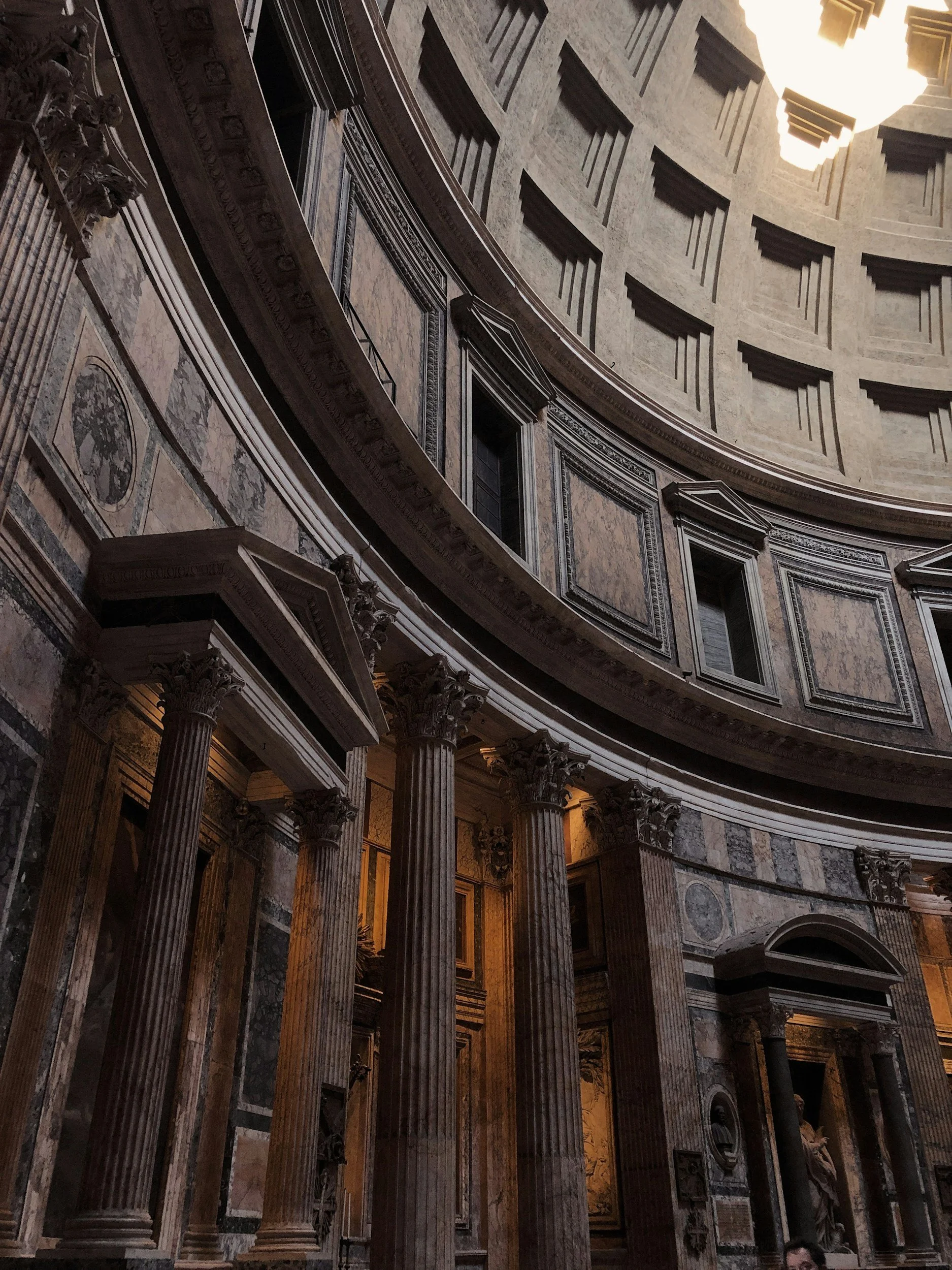 Interior of a grand historic building with large marble columns, ornate decorative details, and an oculus ceiling with a bright light shining through.