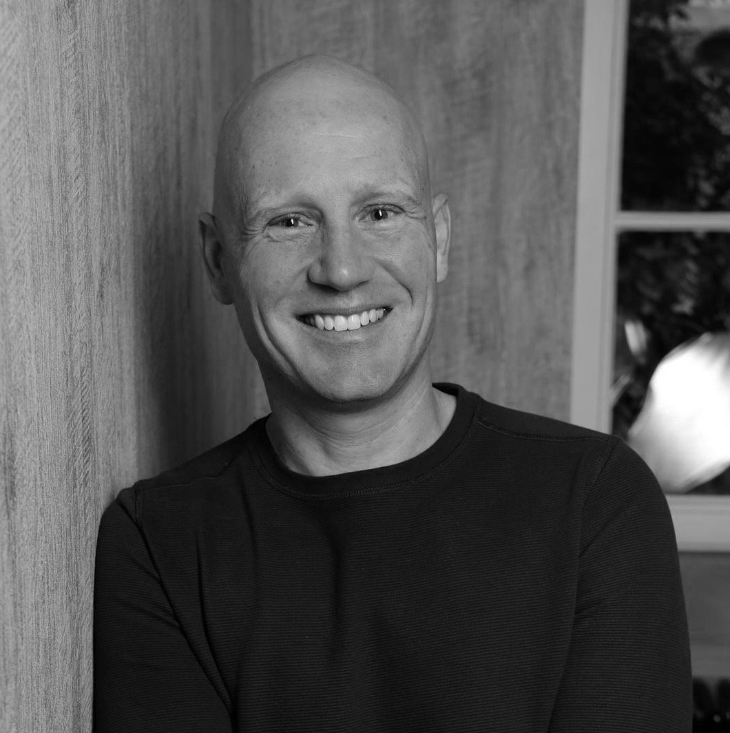Black and white photo of smiling man with a shaved head, wearing a dark shirt, standing against a wooden wall near a window.