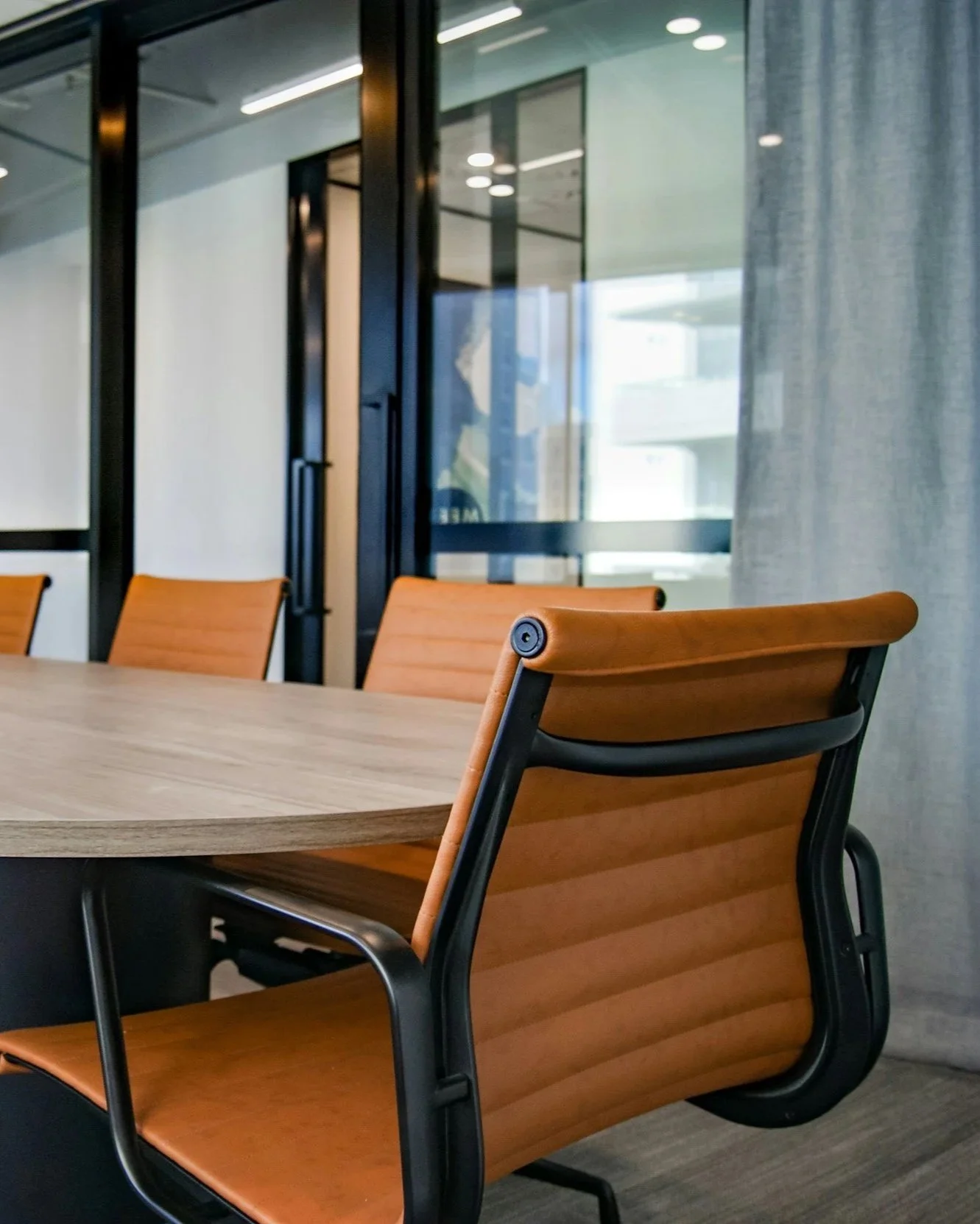 Empty conference room with wooden chairs, a round table, glass walls, and gray curtains.