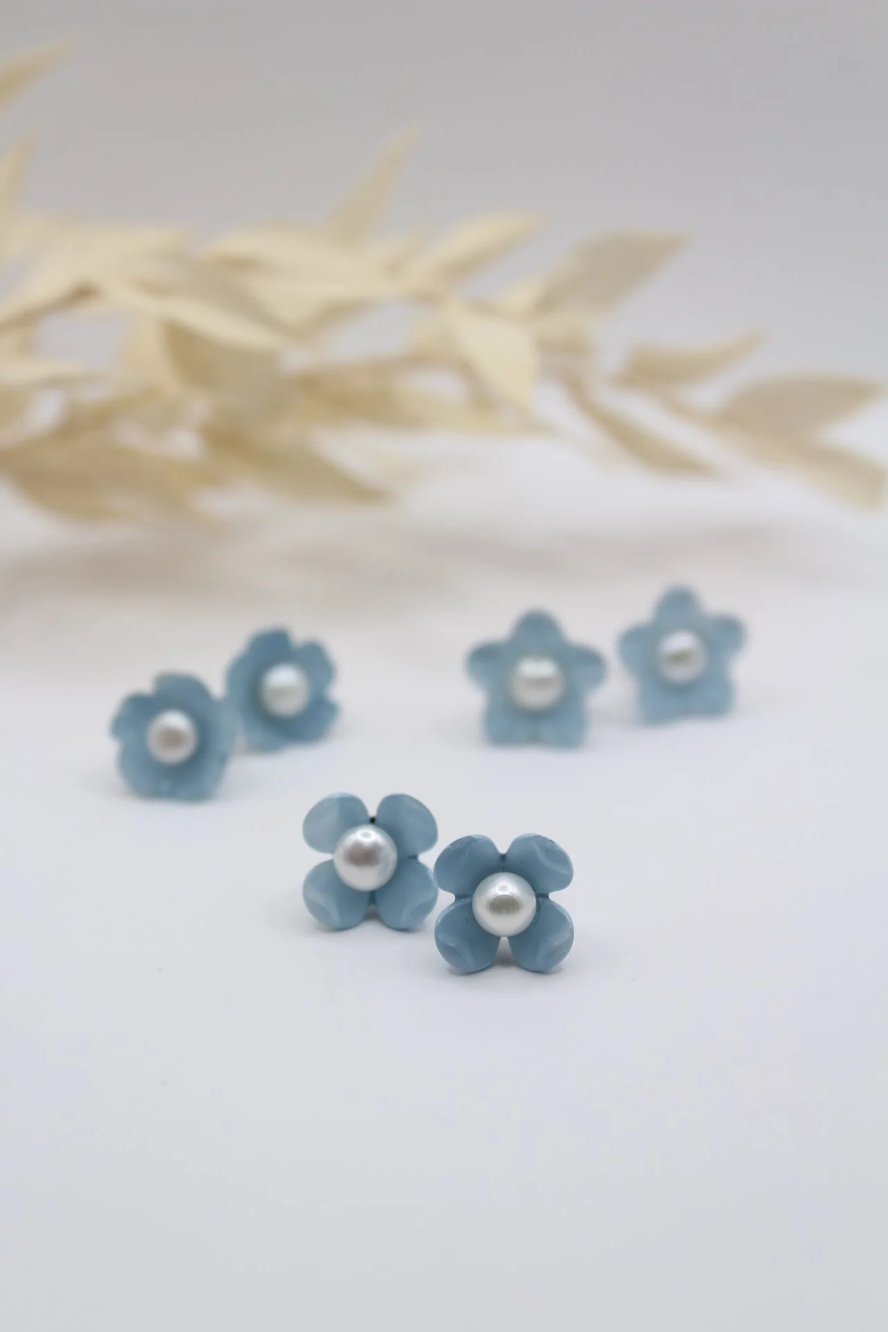Vier blaue Blumen-Ohrringe mit Perlen in der Mitte, leuchten vor einem weißen Hintergrund.