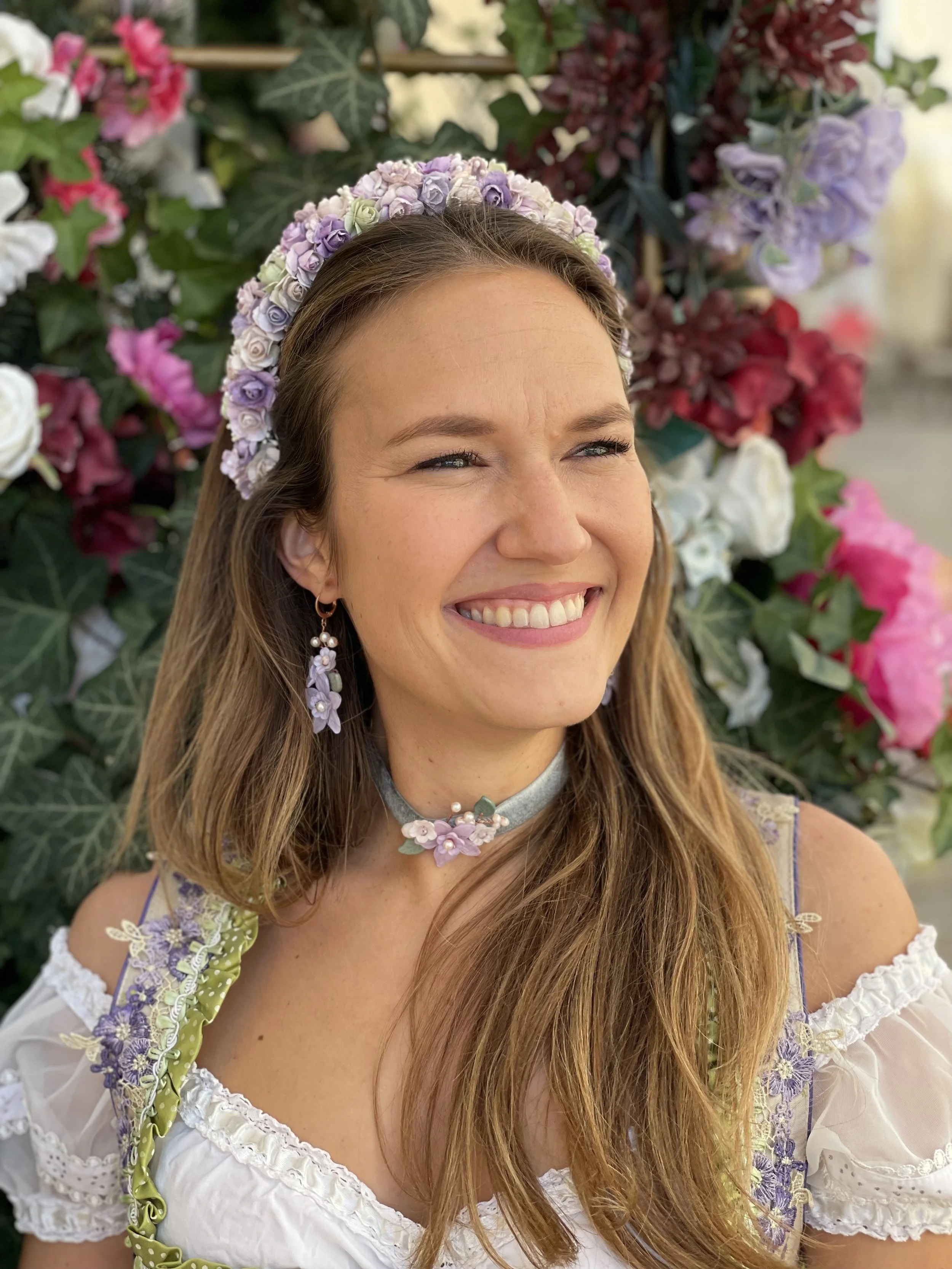 Eine lächelnde junge Frau mit Blumenkranz auf dem Kopf und Blumenohrringen, umgeben von bunten Blumen im Hintergrund.