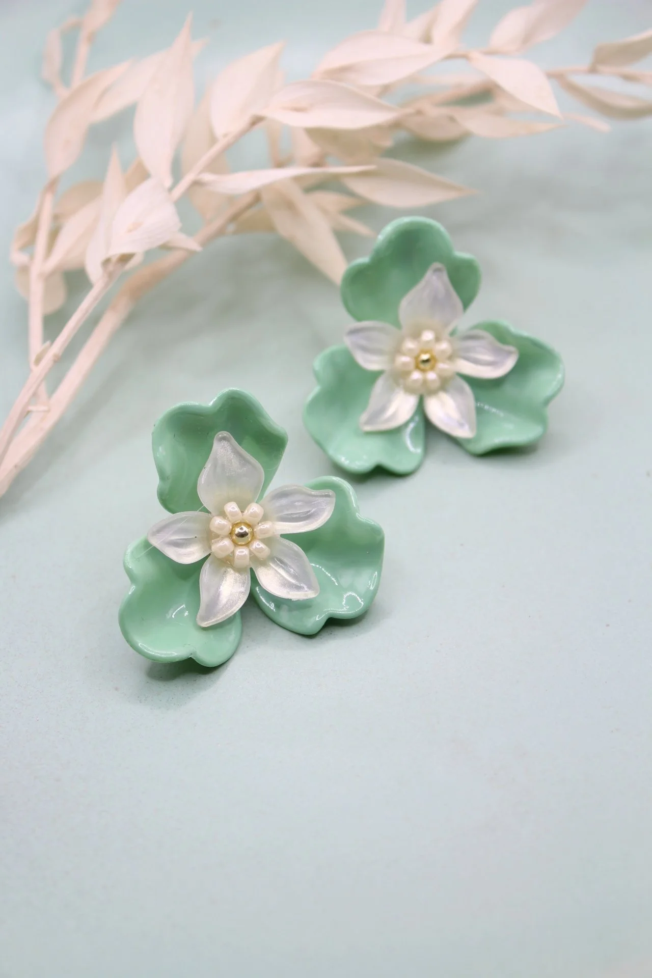 Zwei grüne, blumenförmige Ohrringe mit weißen Blüten und Perlen in der Mitte, auf einem hellblauen Untergrund mit einer weißen Zweig-Deko im Hintergrund.