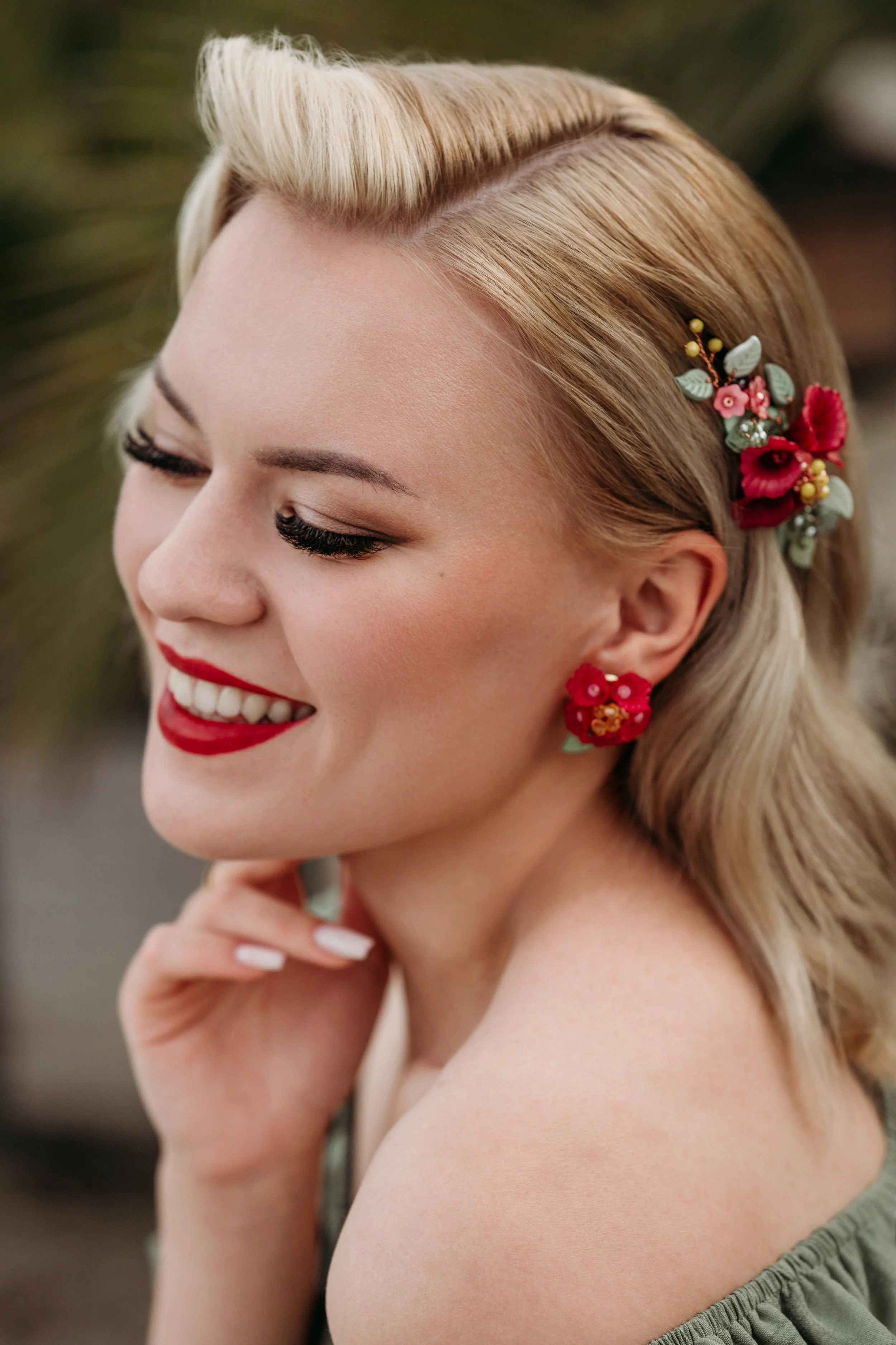 Ein lächelndes Mädchen mit blondem Haarschmuck und roten Blumenohrringen, das die Augen geschlossen hat.