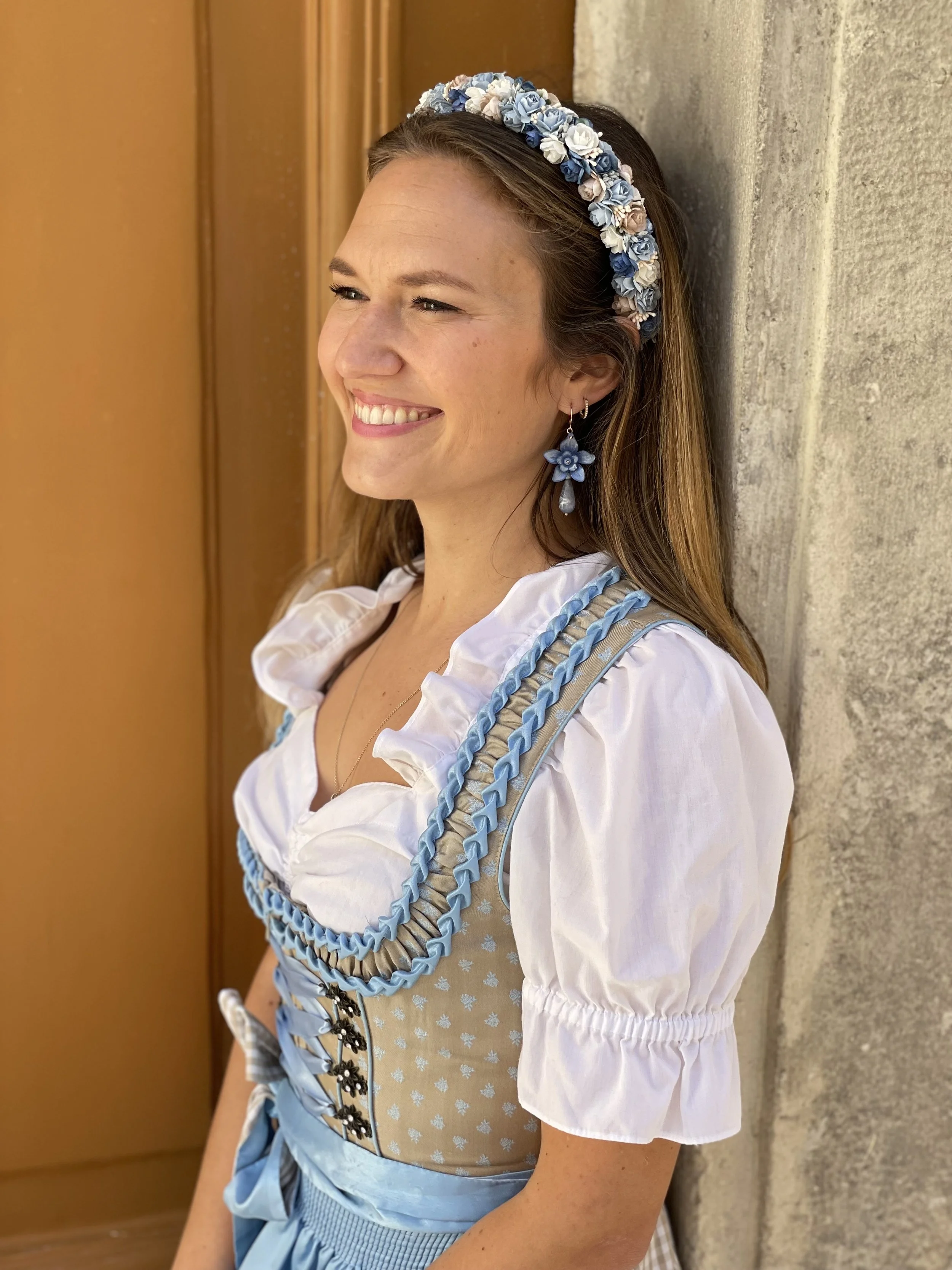 Eine lachende Frau im traditionellen bayerischen Dirndl mit blumenverziertem Haarband und Ohrringen, die an einer Mauer lehnt.