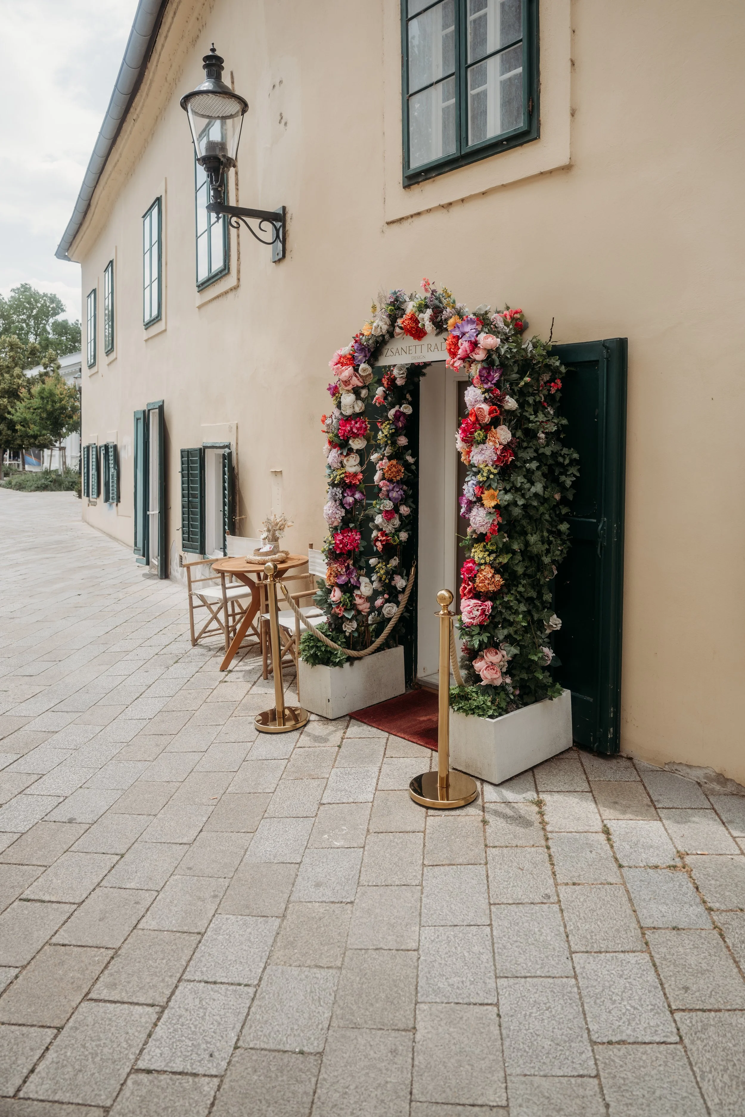 F negatively decorated Eingang mit Blumen und goldenen Absperrpfosten in der Nähe eines Gebäudes mit weißen Wänden und grünen Fensterläden.