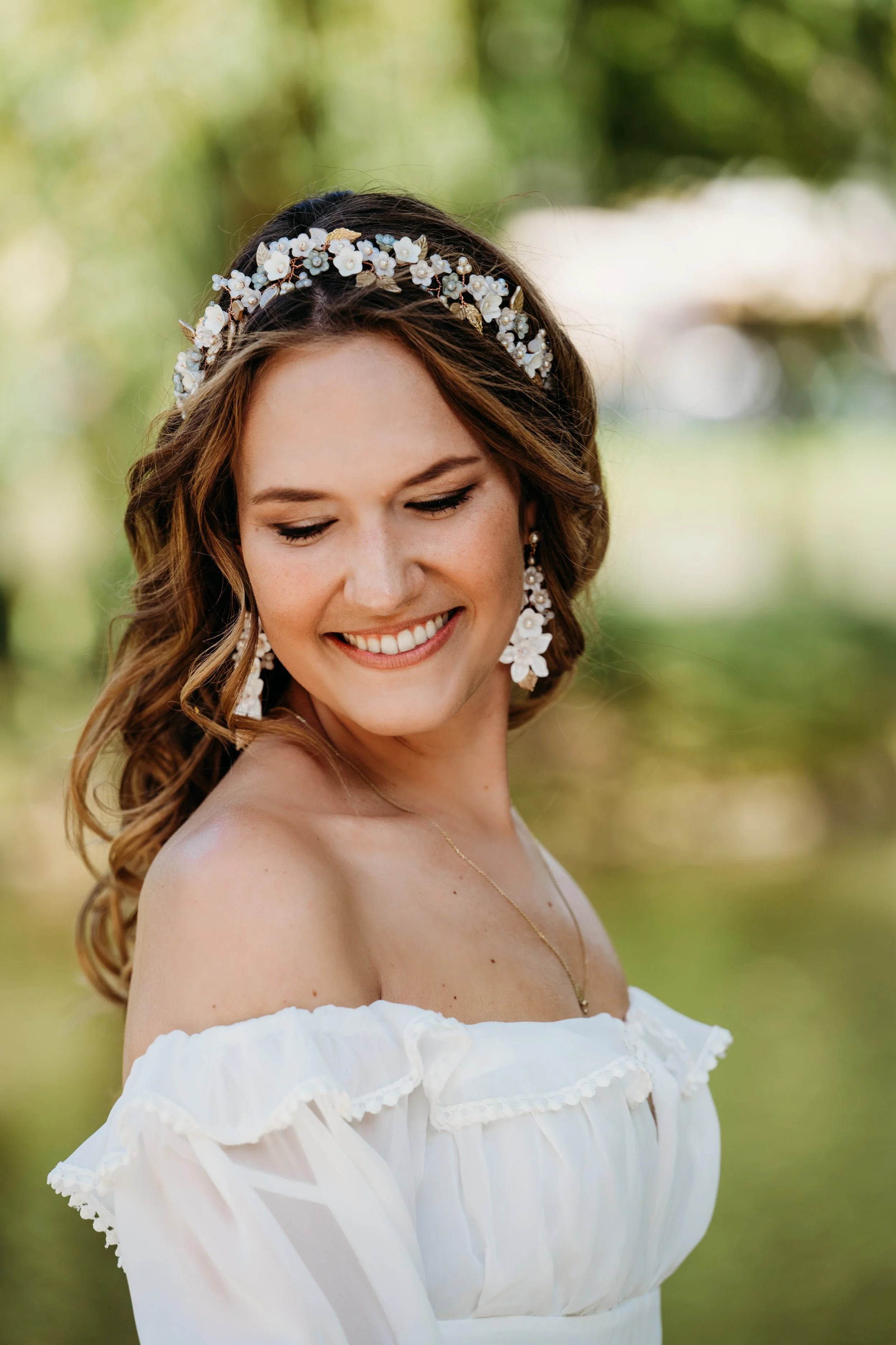 Eine lächelnde Frau in einem weißen Kleid mit Blumenverzierung und Ohrringen, im Freien, umgeben von grünen Bäumen.