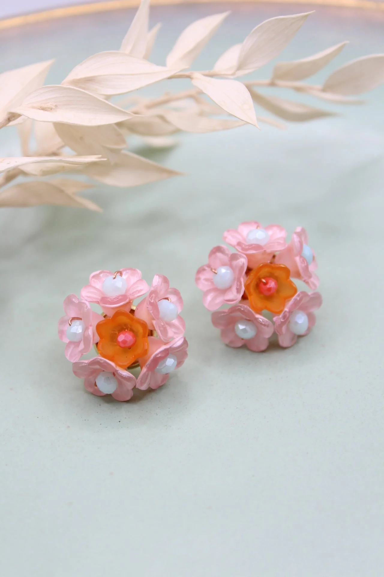 Zwei Perlen- und Kunststoffblumenohrringe in Pink und Orange, auf einem hellen Hintergrund. Oben ein gestreifter Zweig.