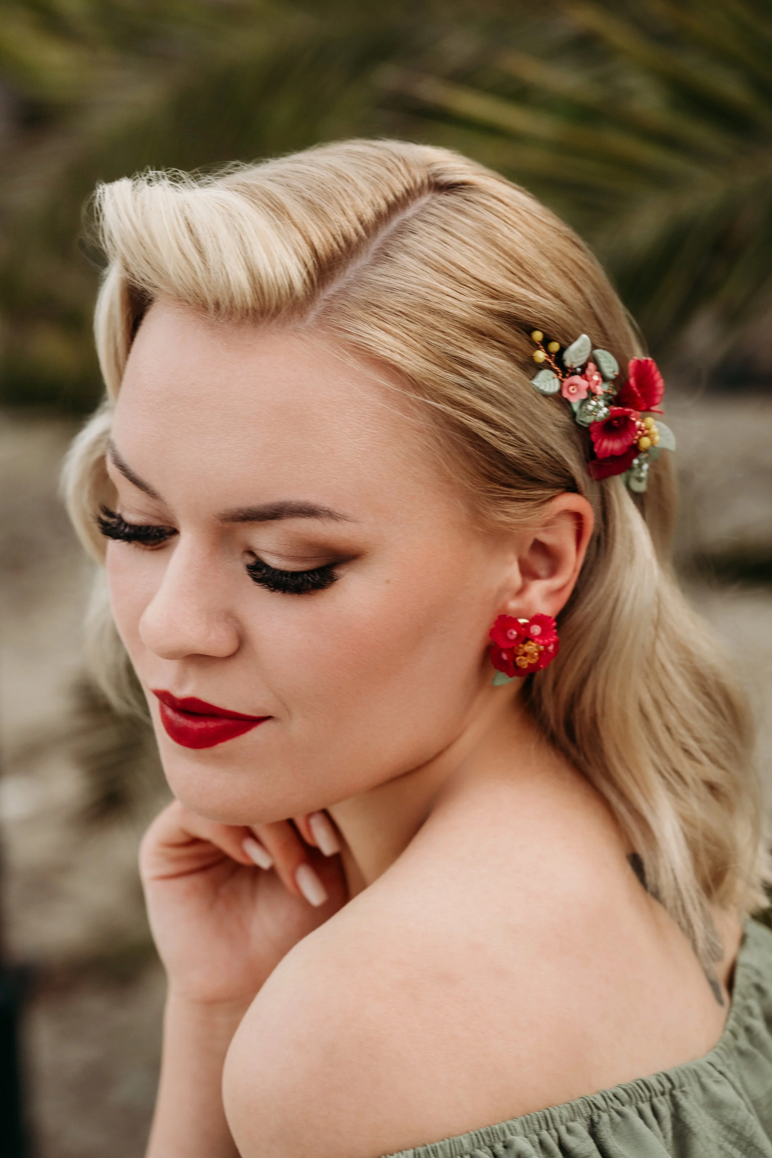 Nahaufnahme eines weiblichen portraits mit blondem haare, rotem lippenstift, rotem blumenohrring und blumen im haar, im freiluft hintergrund