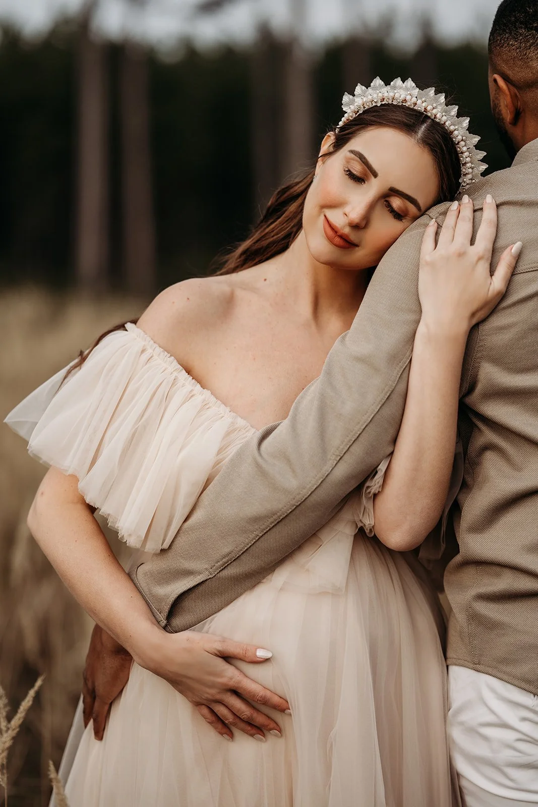 Ein Paar umarmt sich in einem Feld, die Braut trägt ein cremefarbenes off-shoulder Kleid mit Tüllfalten und ein Blumenkronenkopf.