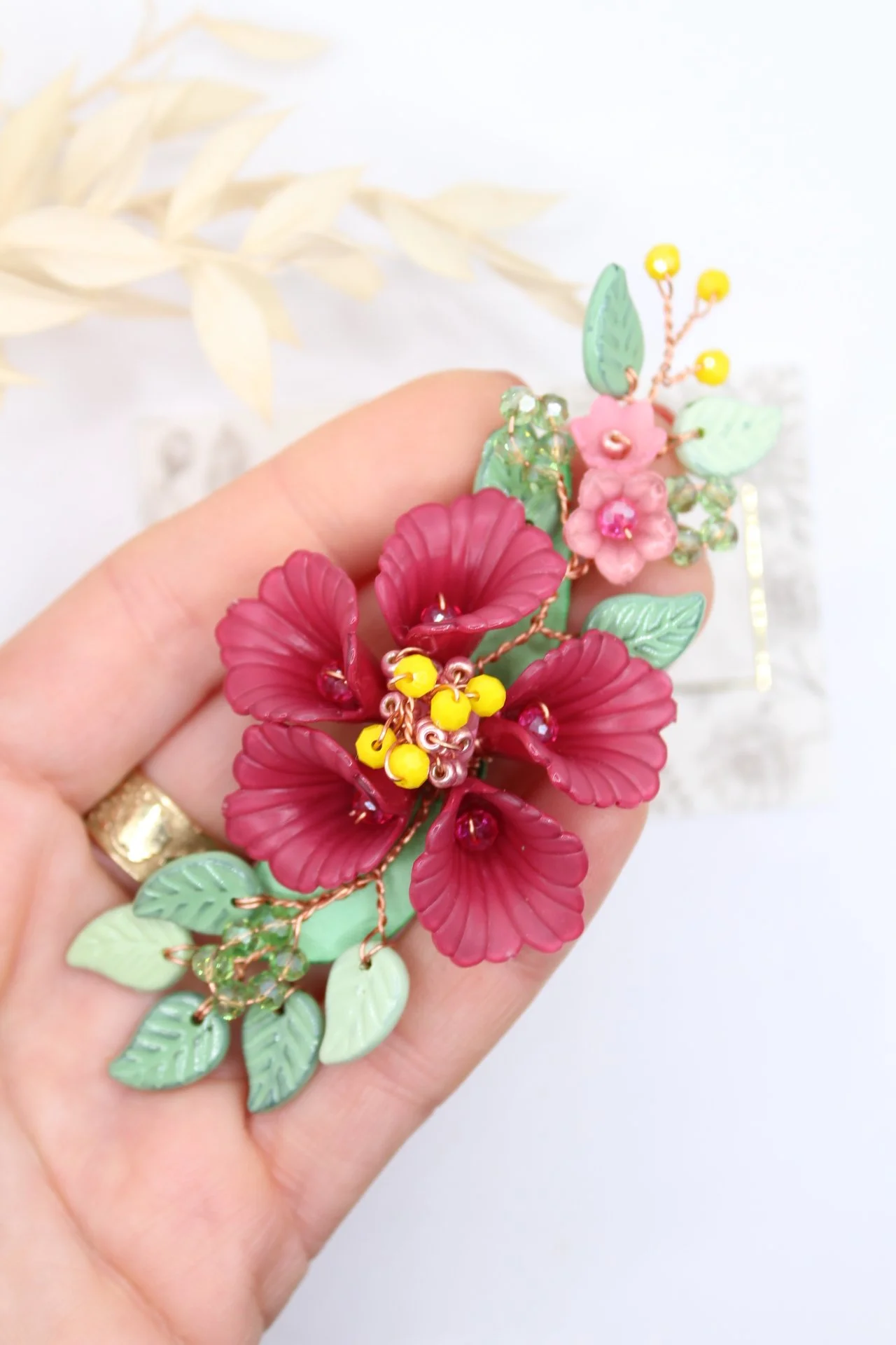 Hand hält eine bunte Halskette mit roten Blüten, grünen Blättern, gelben und pinken Akzenten, und kleinen Schmuckelementen, vor einem weißen Hintergrund.