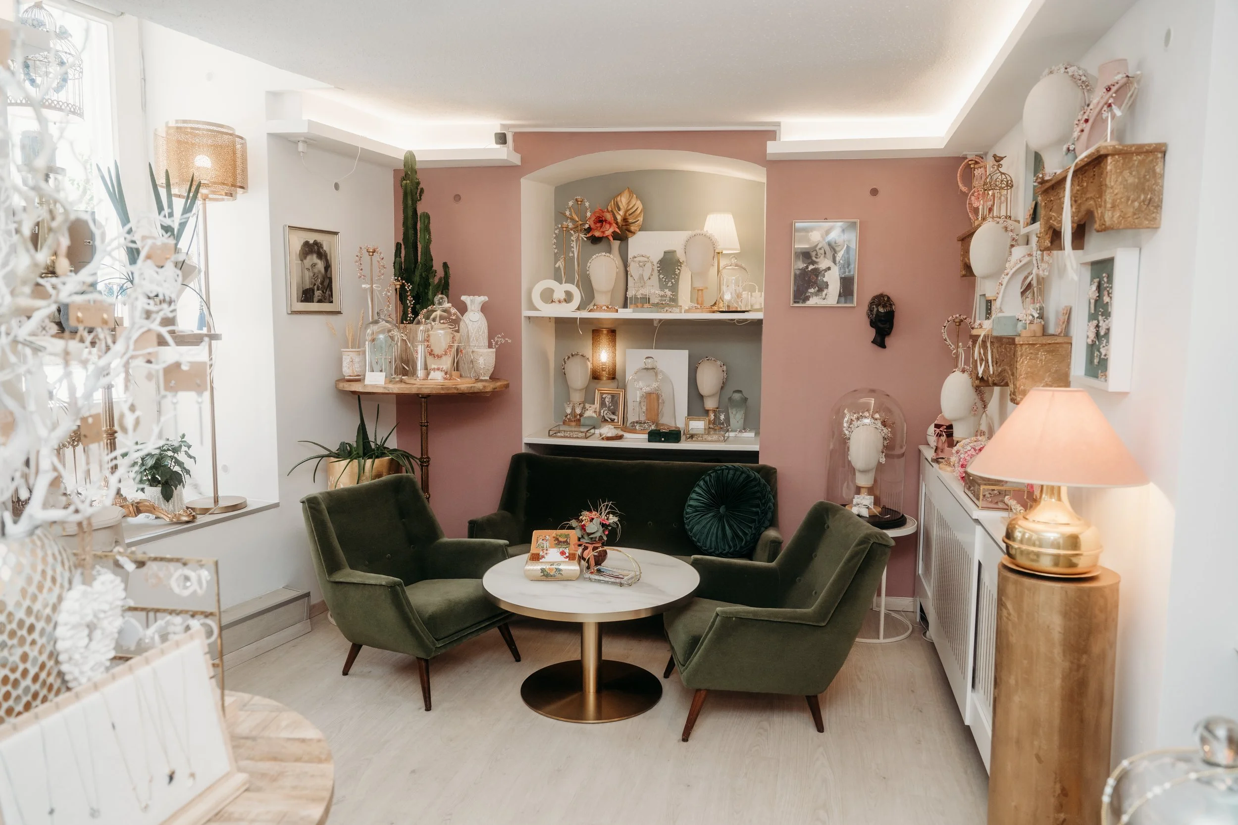 Gesamtszene eines Verkaufsraums mit schmuck und Dekorationsartikeln, grüner couch, runder Tisch, pinke und weiße Wände, dekorative Regale, Lampen und Pflanzen.