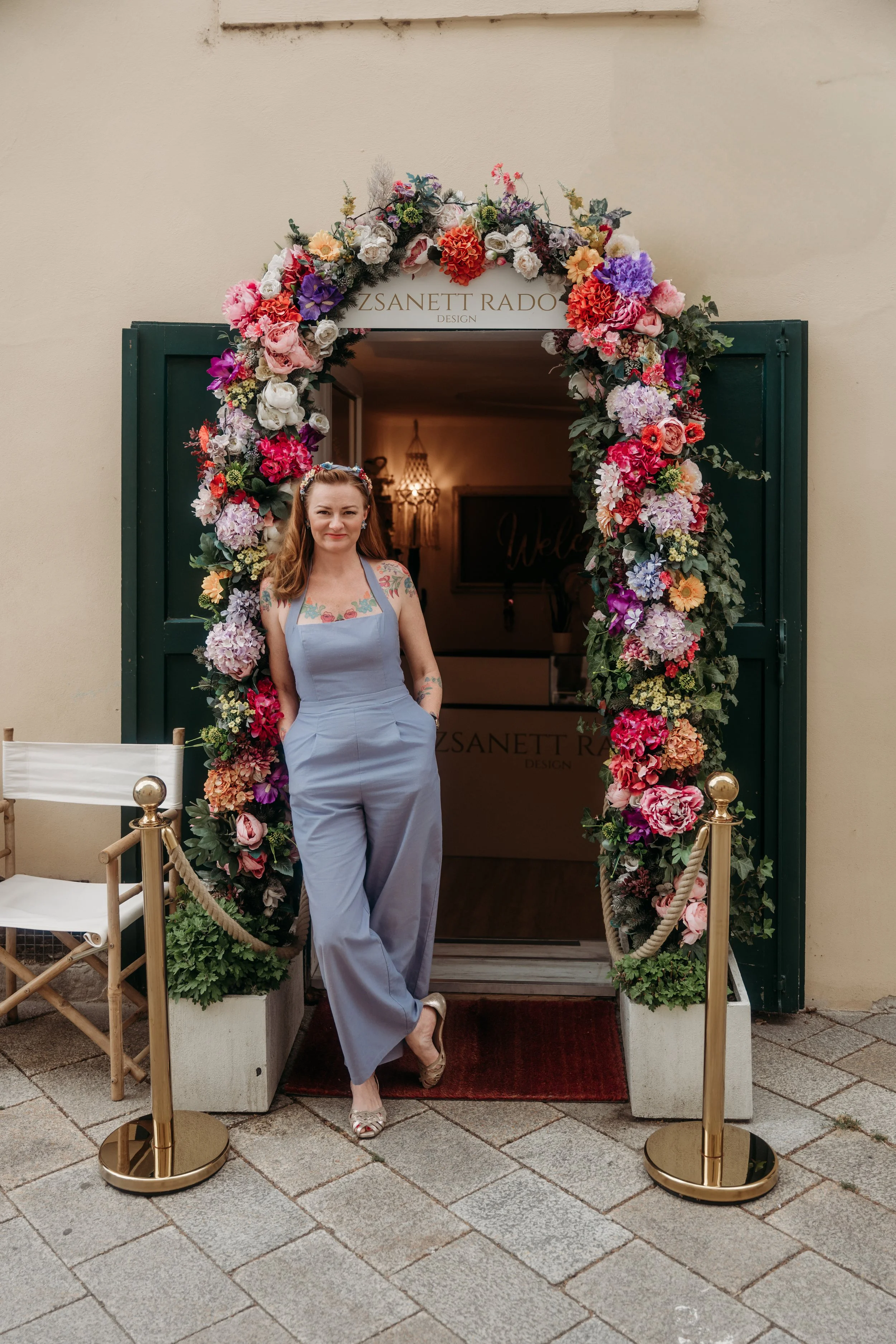 Eine Frau steht vor einem Eingangsbereich, der von einem blumengeschmückten Bogen eingerahmt wird. Sie trägt einen hellblauen Jumpsuit und Schuhe, und hat lange, lockige Haare.