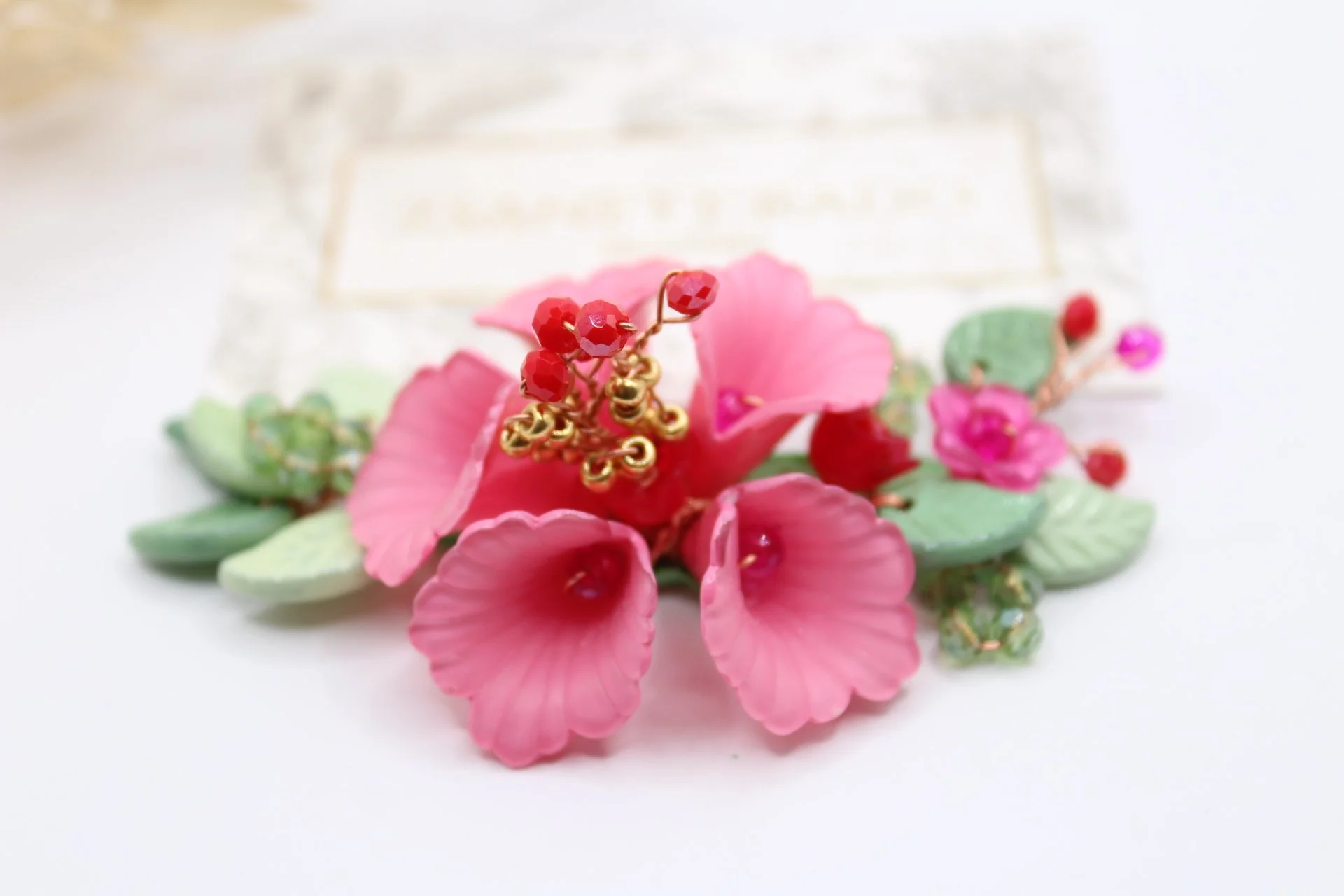 Nahaufnahme eines pinken Blumenbroschen mit grünen Blättern und roten Perlen auf weißem Hintergrund.