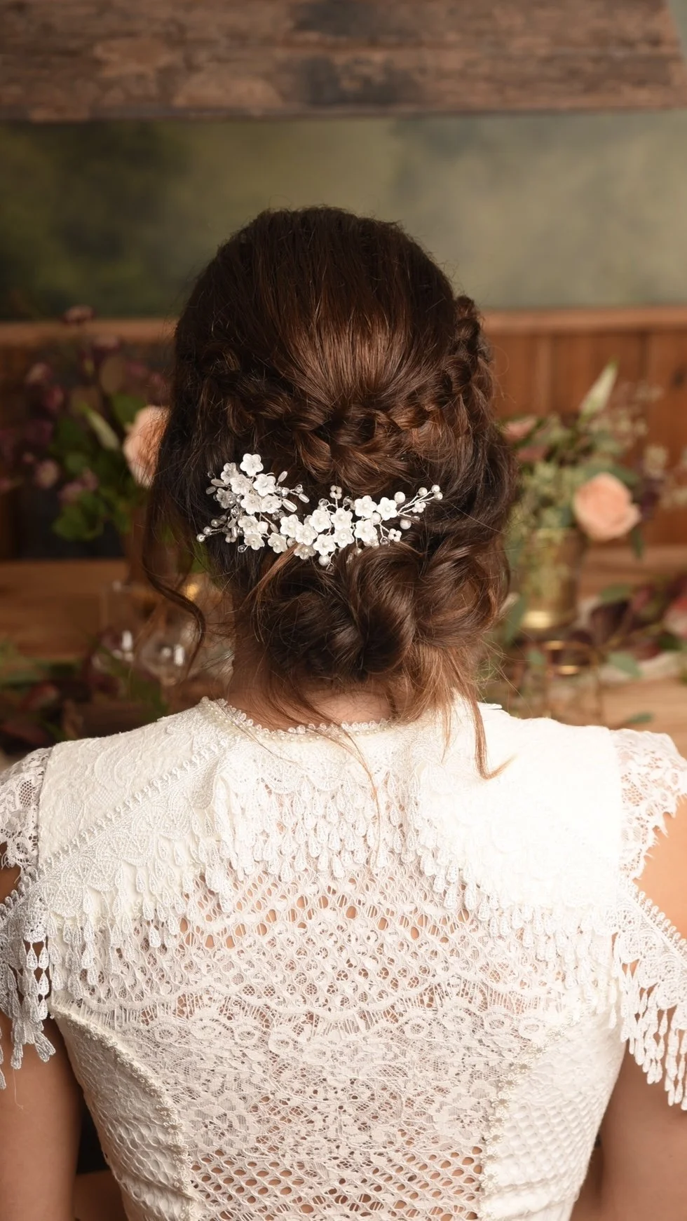 Rückansicht einer Frau mit braunen, lockigen Haaren, die zu einem Chignon gestylt sind, mit einer weißen Haarblume und Perlen, auf einer Hochzeit oder festlichen Veranstaltung, im Hintergrund Blumen und eine Holzwand.