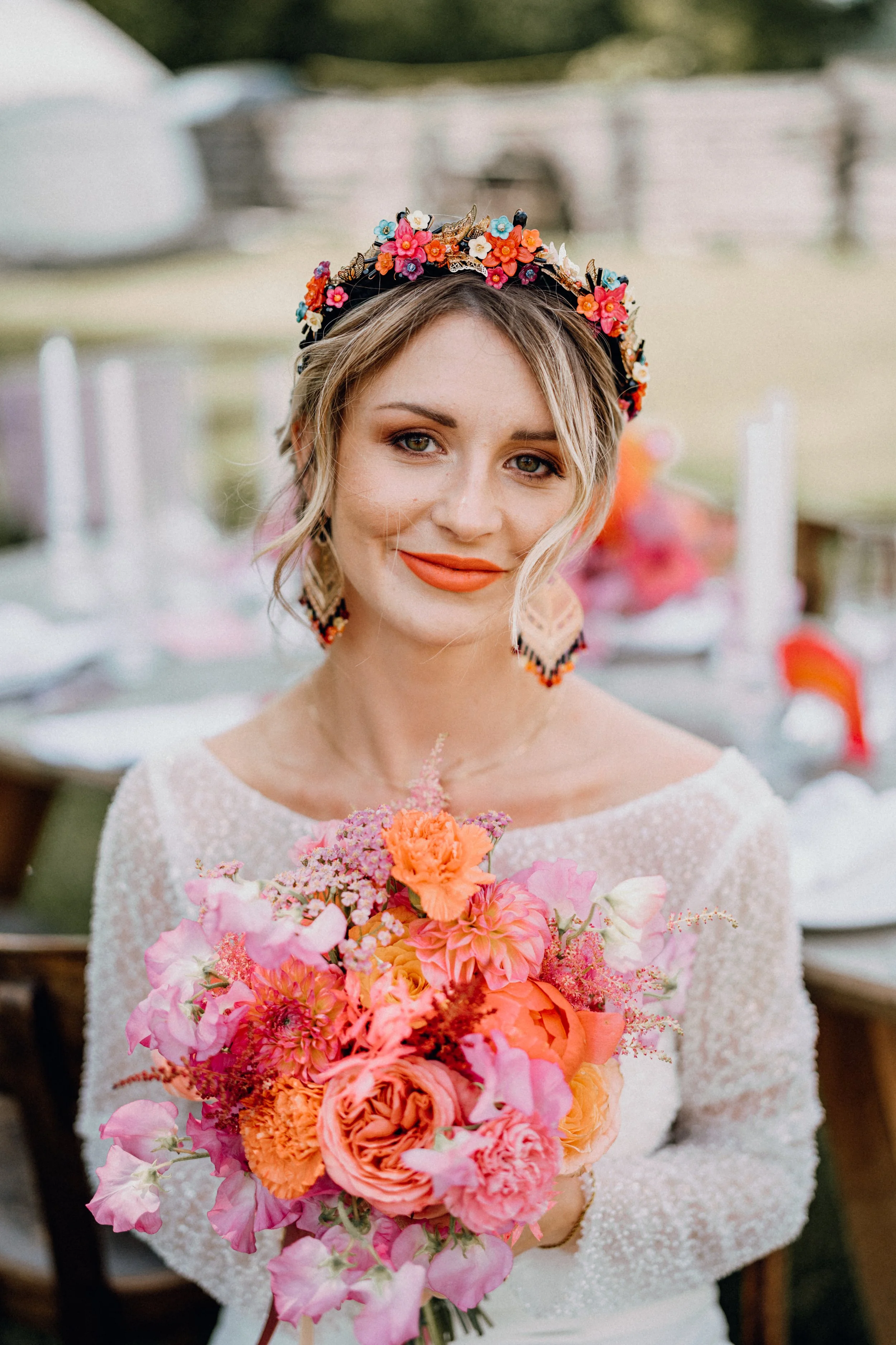 Junge Frau mit blumengeschmücktem Kopf und großem Blumenstrauß bei einer Hochzeit im Freien, lachend.