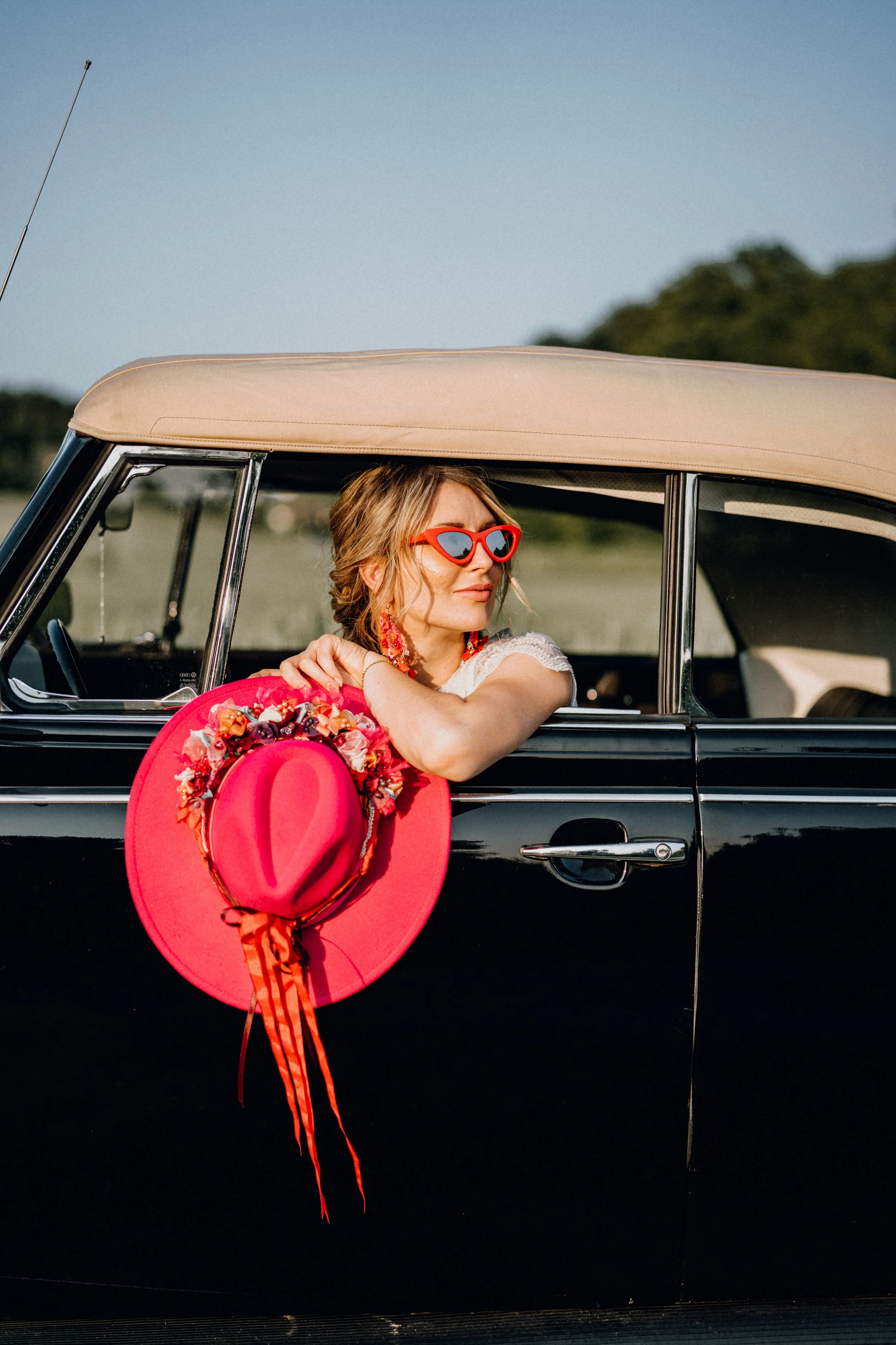 Frau mit roter Sonnenbrille, roten Ohrringen und einem roten Hut mit Blumen, lehnt an einem offenen Autofenster in einem schwarzen Cabriolet, im Freien bei Sonnenschein.