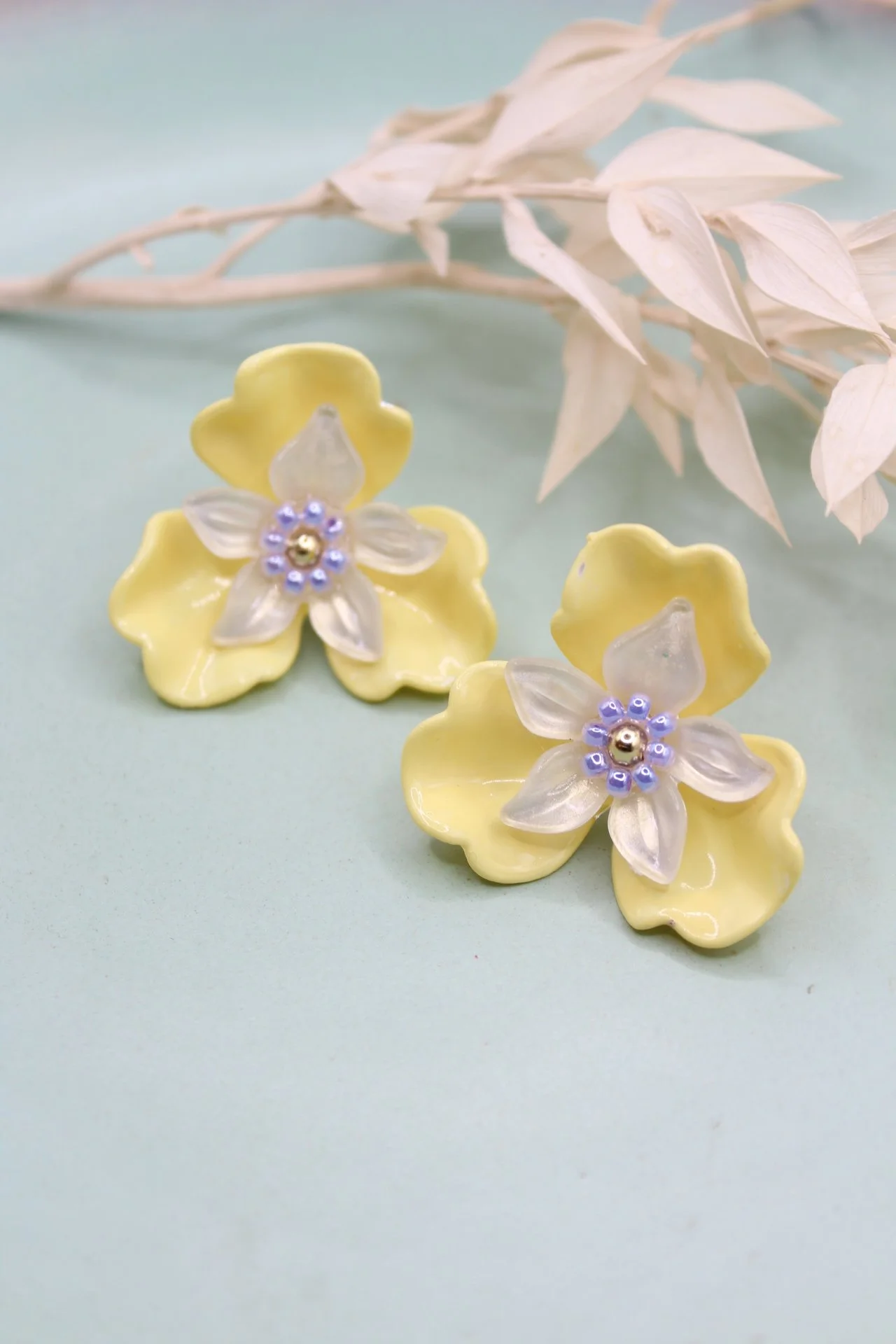 Gelbe Blumenohrringe mit weißen Blüten und blauen Perlen, daneben weiße getrocknete Blätter auf einem grünen Hintergrund.