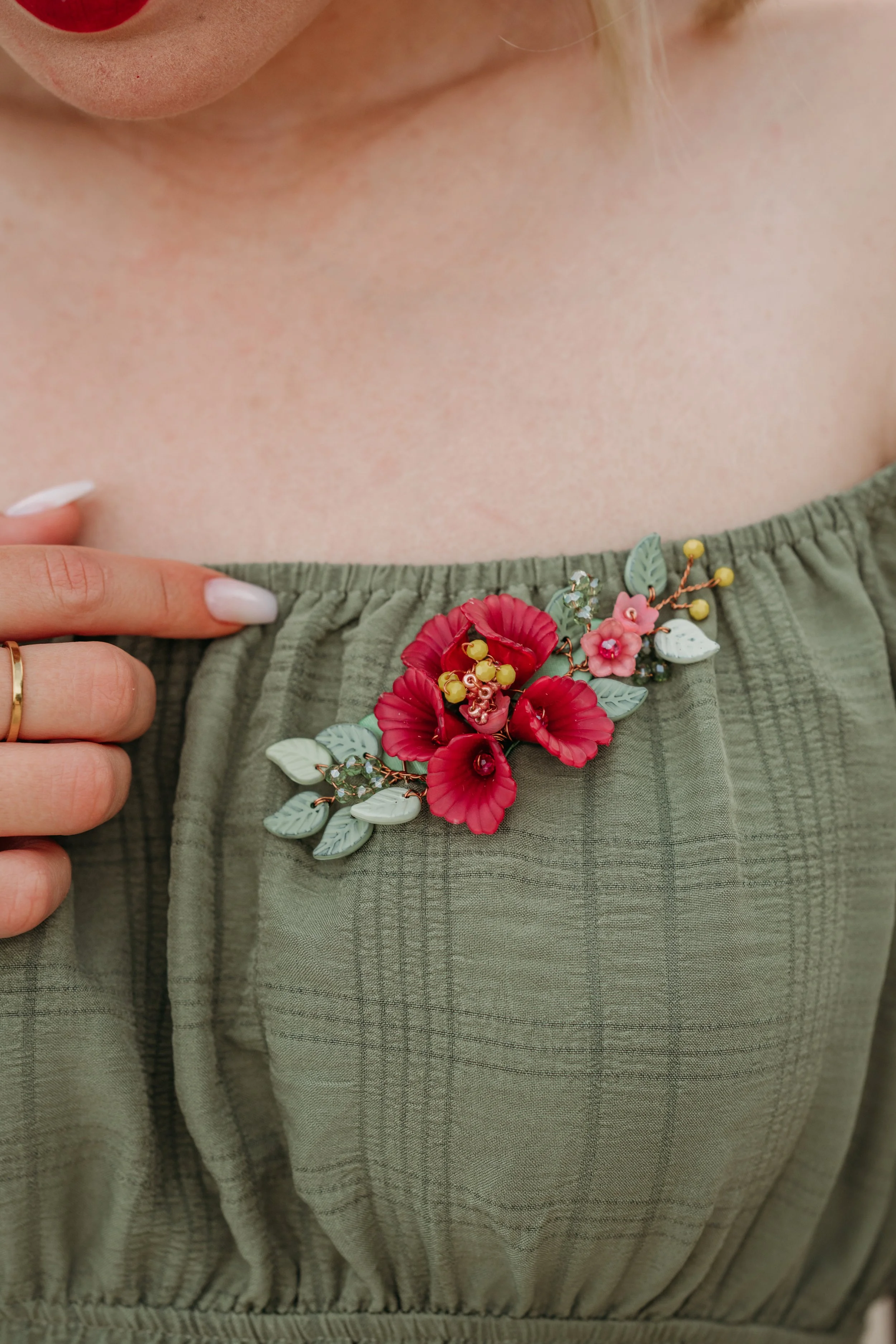 Nahaufnahme eines grünen Kleides mit einer großen, roten Blumenbrosche und einem Finger mit Goldring.