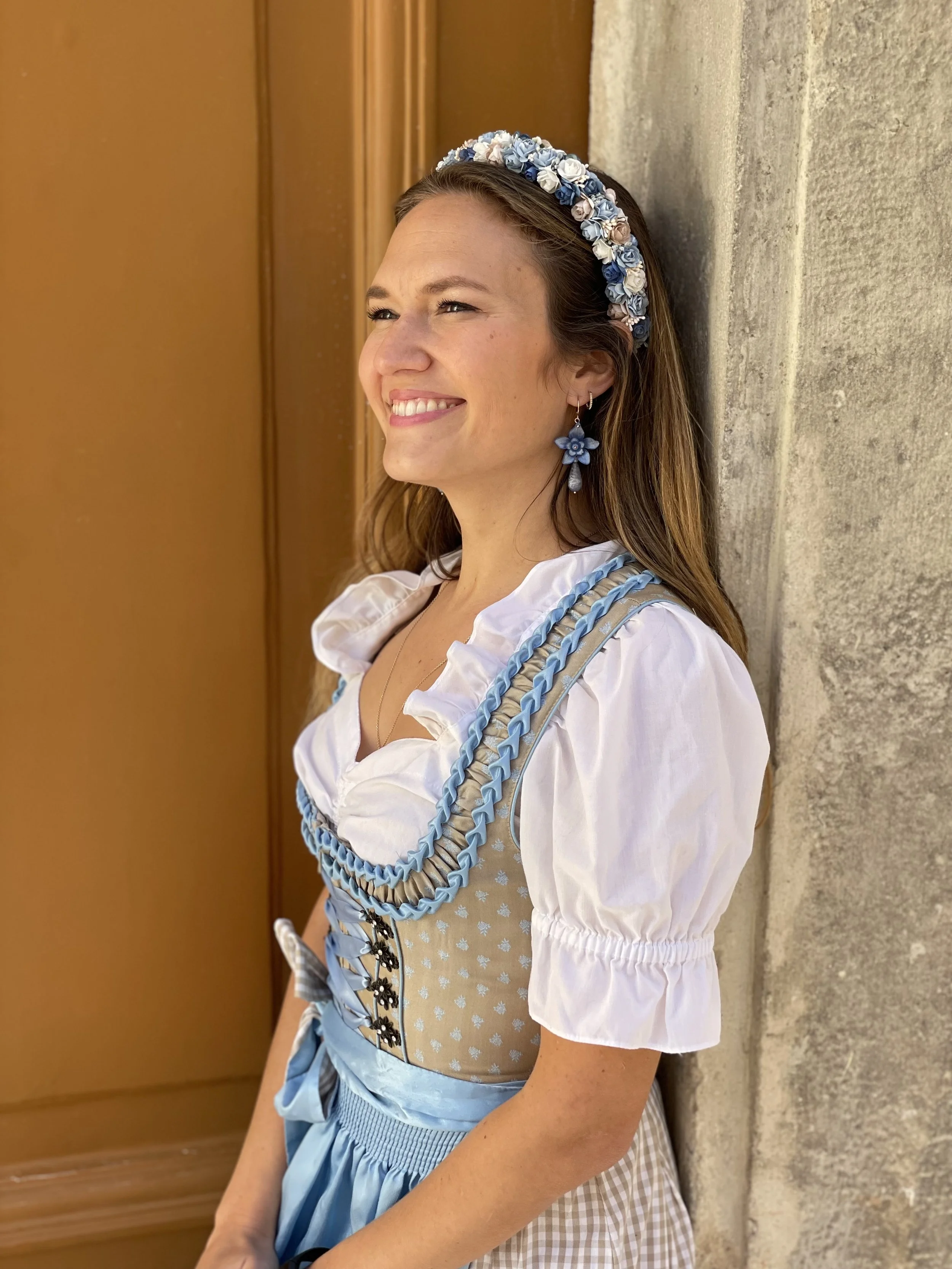 Junge Frau in traditioneller bayerischer Tracht mit lavendelfarbener Bluse, beige Mieder mit blauen Zierkordeln, kariertem Rock und floralem Haarschmuck, die an einer Steinmauer lehnt, lächelnd ins Licht schauend.