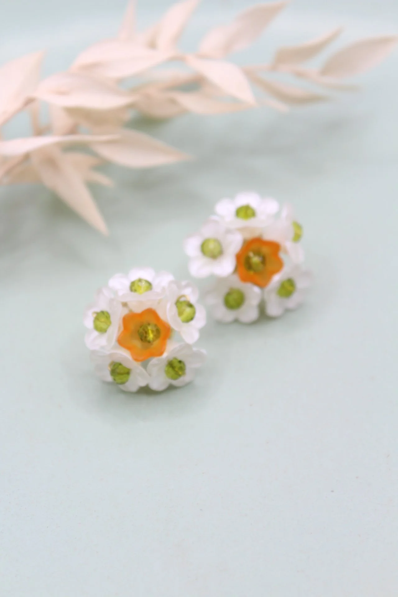 Zwei kleine, bunte Blumenohrringe mit weißen, orangefarbenen und grünen Details, auf einem hellen Hintergrund mit weißen getrockneten Blättern im Hintergrund.