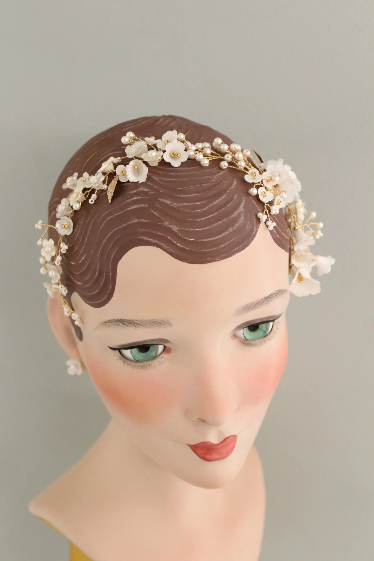 Haarreif mit kleinen weißen Blumen und Perlen auf einer weiblichen Modepuppe mit grünen Augen und rotem Lippen. Hintergrund grau.