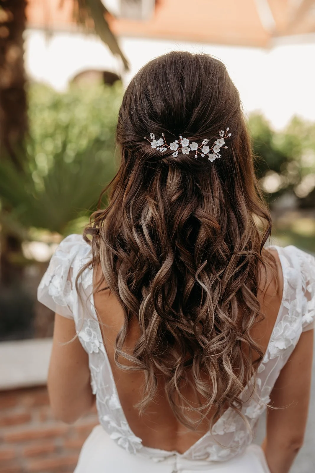 Eine Frau mit lockigem braunem Haar, die eine weiße Bluse trägt und eine silberne Haarschmuck in Form von Blumen hat, ist von hinten fotografiert. Sie steht im Freien mit grünen Pflanzen im Hintergrund.