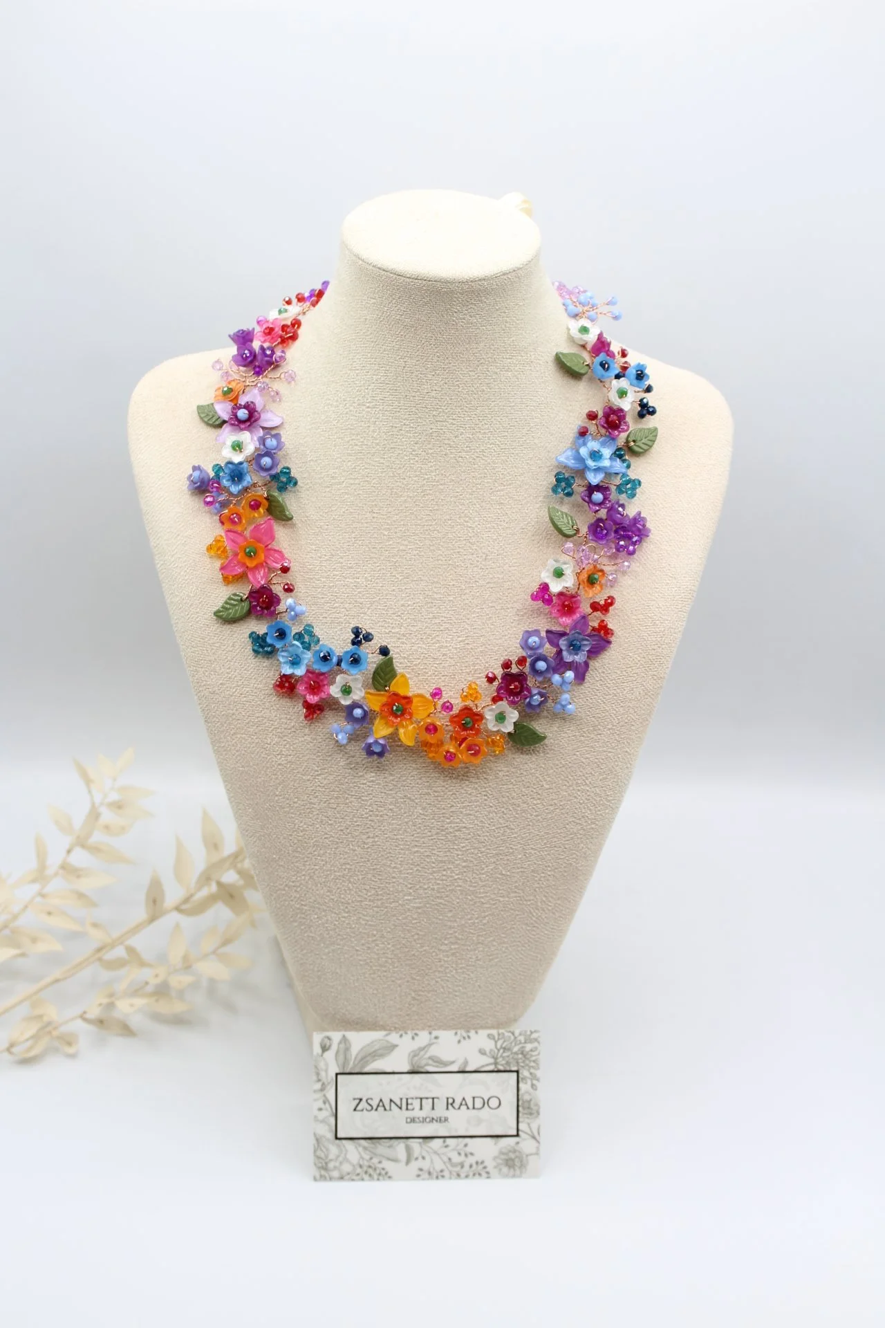 Halskette aus bunten Blumen und Blättern, drapiert um einen Puderrosa Hals. Präsentiert auf einem Stoffmodell, mit einem Namensschild von Zsanett Rado.