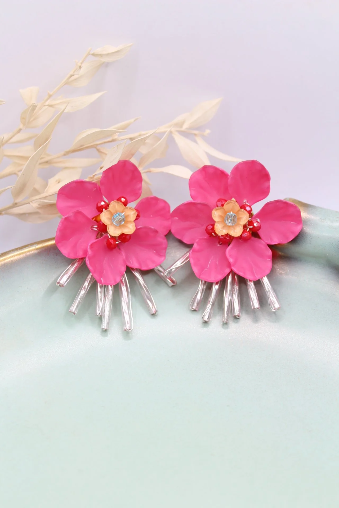 Bunte Blumenohrringe mit pinken Blüten, kleinen roten Perlen, einem blauen Stein in der Mitte und silbernen Stielen, auf einem hellen Untergrund mit weißen Zweigen im Hintergrund.