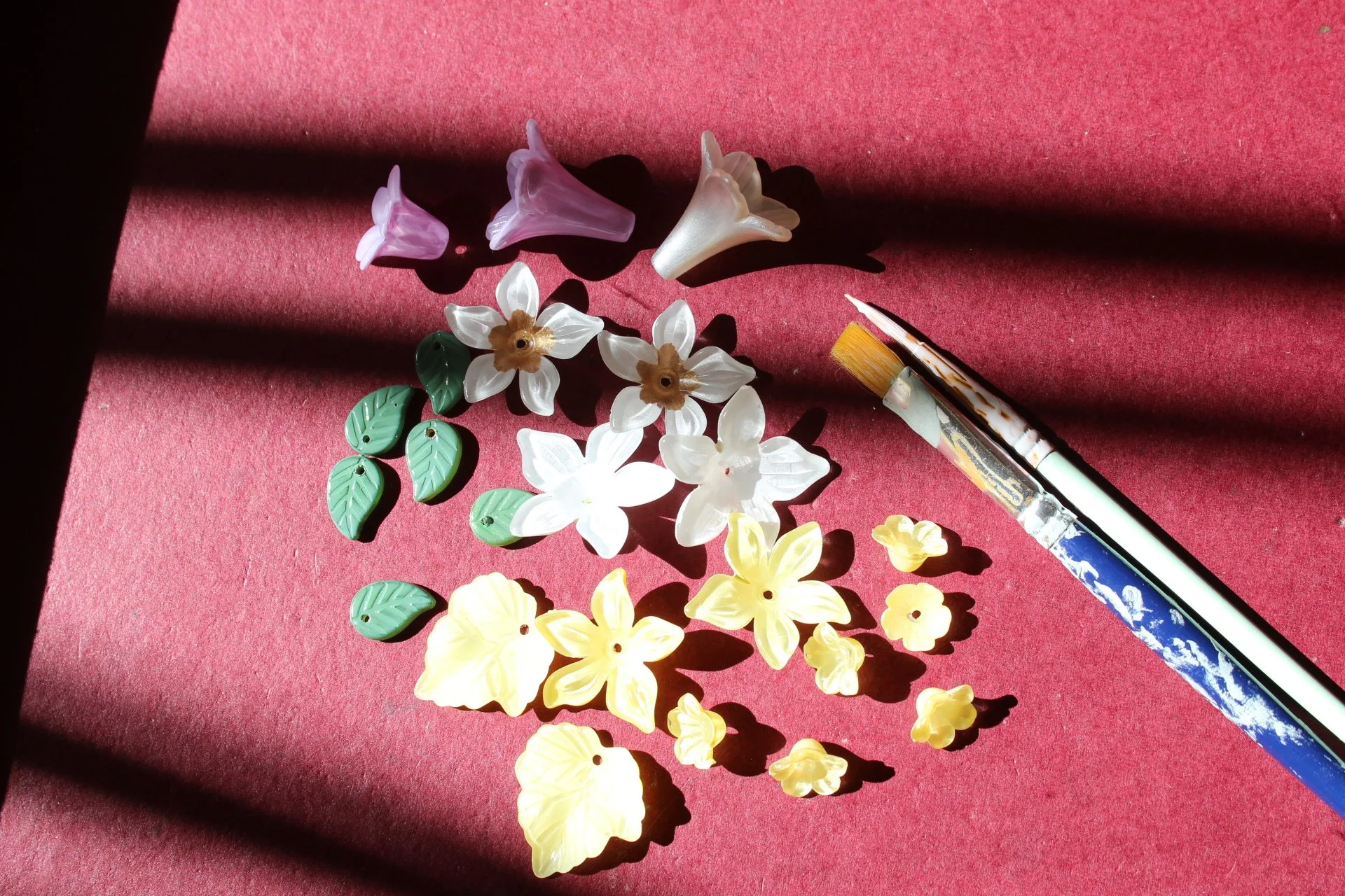 Verschiedene bunte Blumen- und Blätteranhänger aus Kunststoff, ein Pinsel und ein Stift liegen auf rotem Stoff, der Sonnenlichtstrahlen und Schatten wirft.
