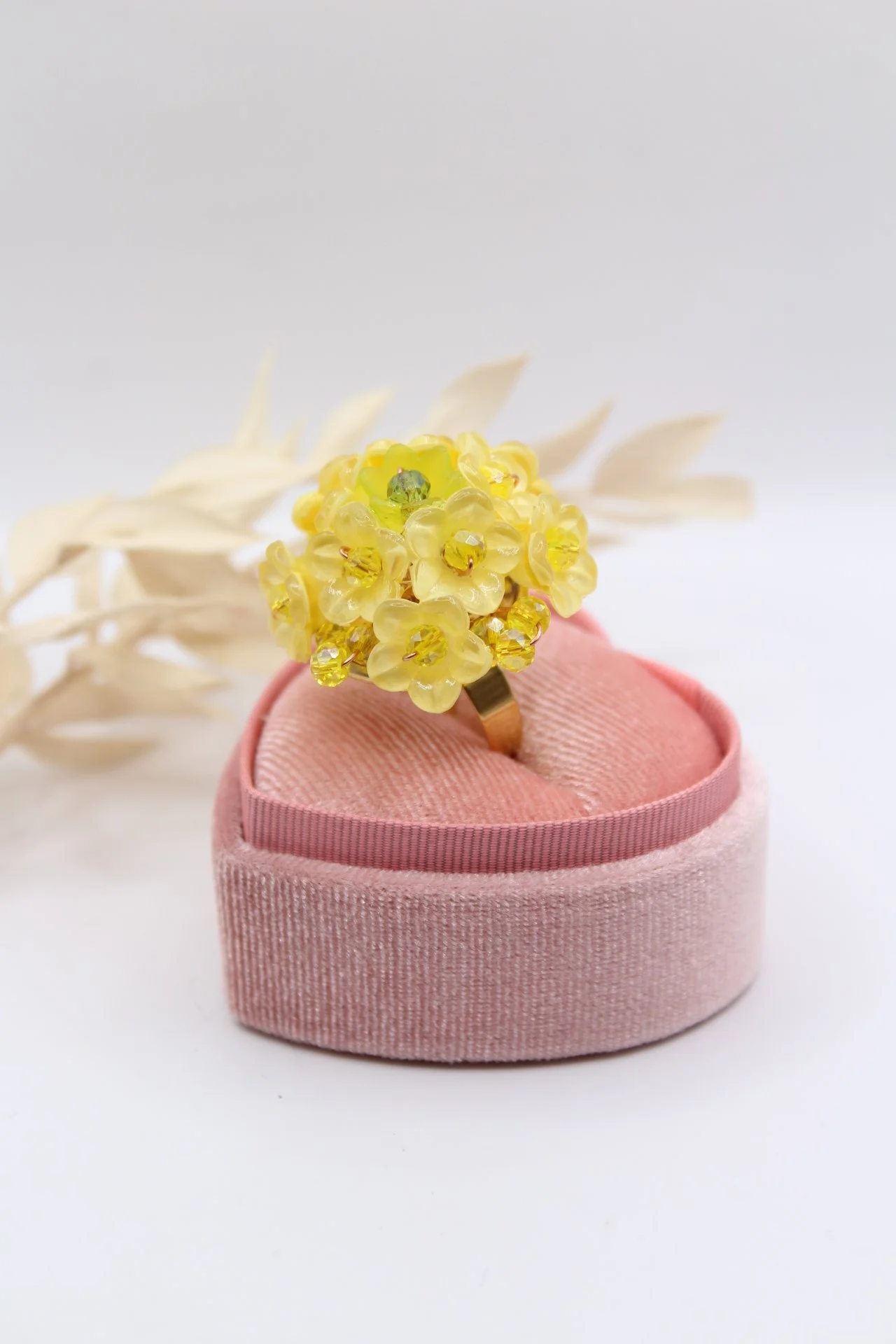 Ein Ring mit gelben Blüten und Kristallakzenten in einer pinken Schmuckschachtel auf weißem Hintergrund, dekoriert mit weißen Zweigen.