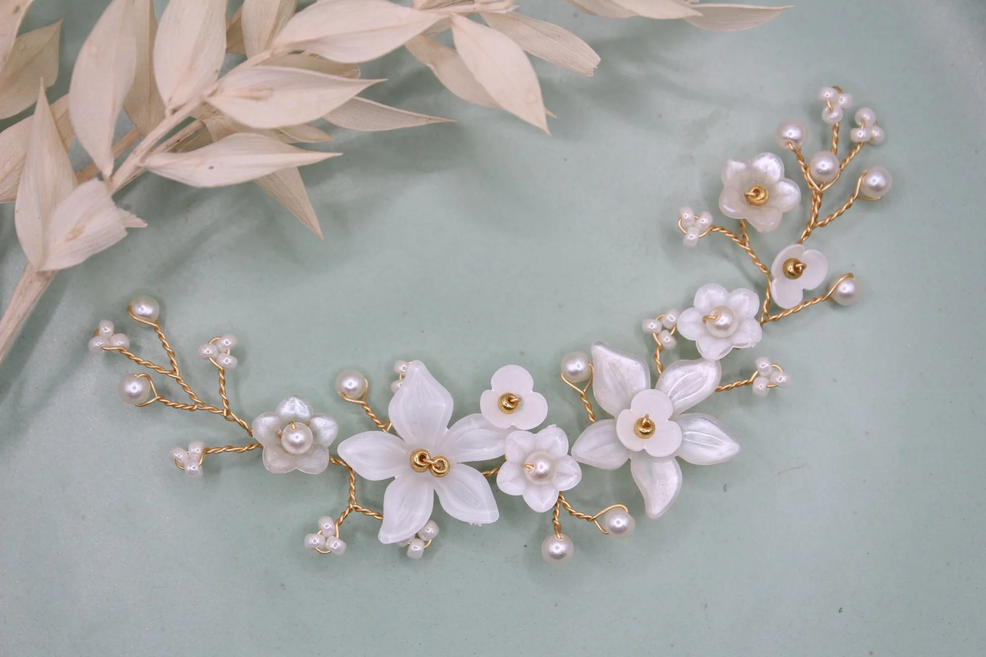 Ein italienisches Haarband mit weißen Blumen und Perlen, auf einem hellgrünen Hintergrund, neben getrockneten weißen Blättern.