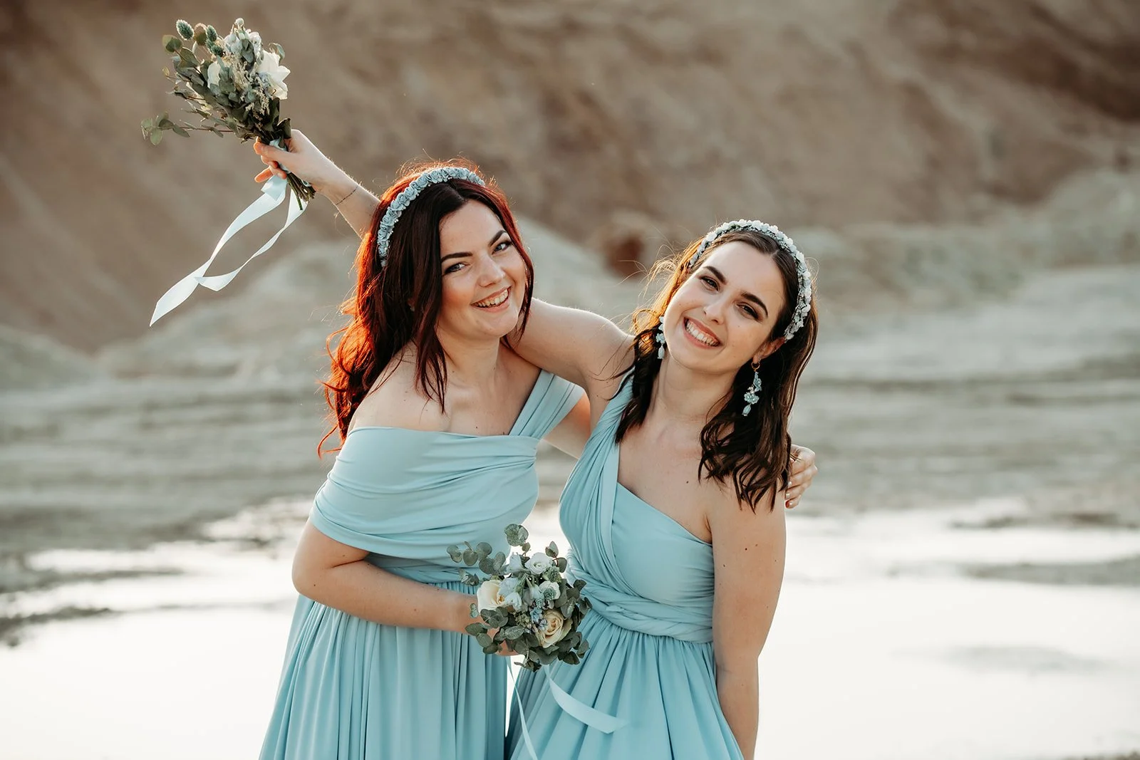 Zwei lachende Frauen in leichten blauen Kleidern mit Blumenaccessoires am Strand, eine hält einen Blumenstrauß hoch.
