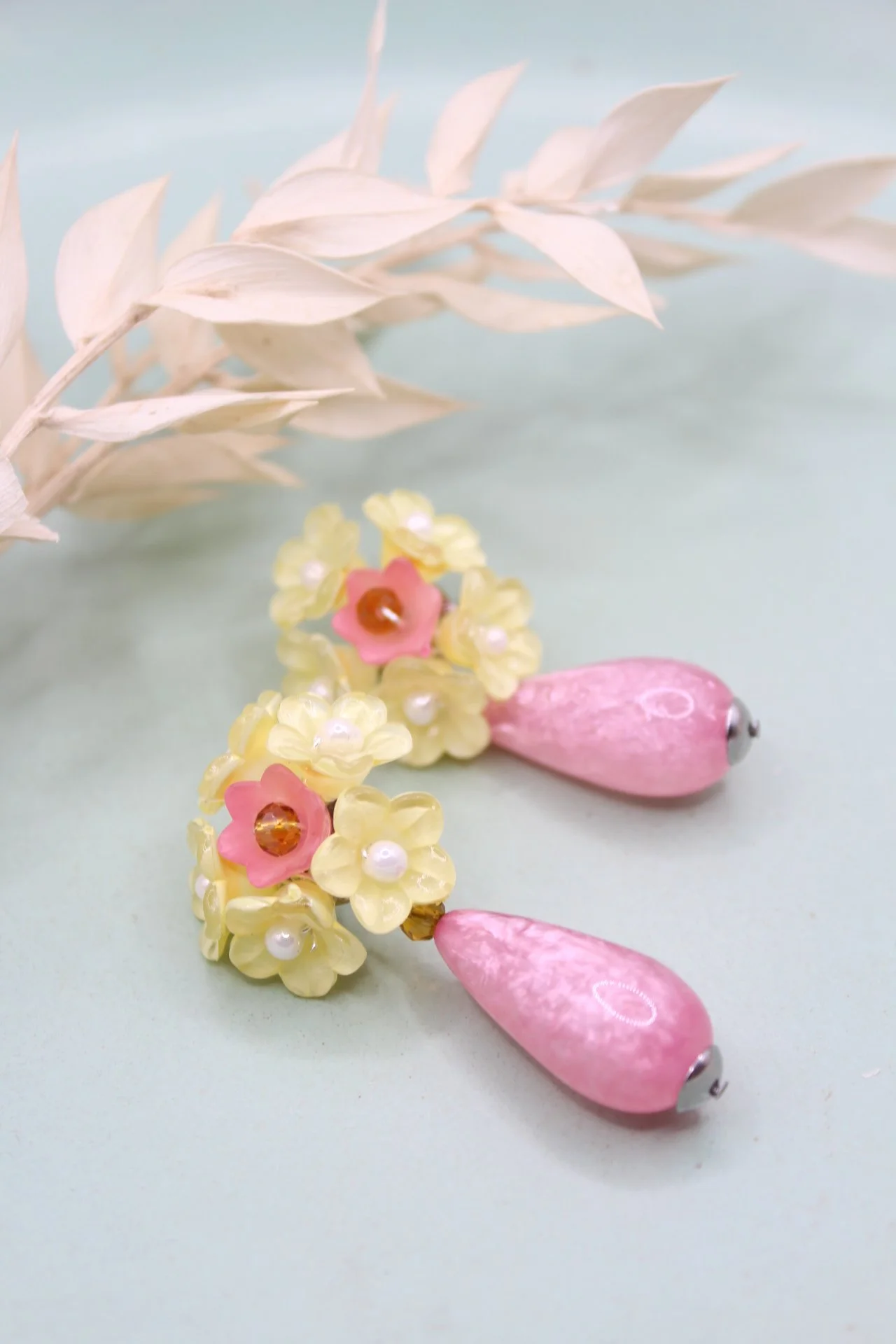 Schmuckstück mit pinken und gelben Perlen, blumenförmigen Elementen und Ziersteinen, daneben eine weiße Blätterzweig auf einem hellen Hintergrund