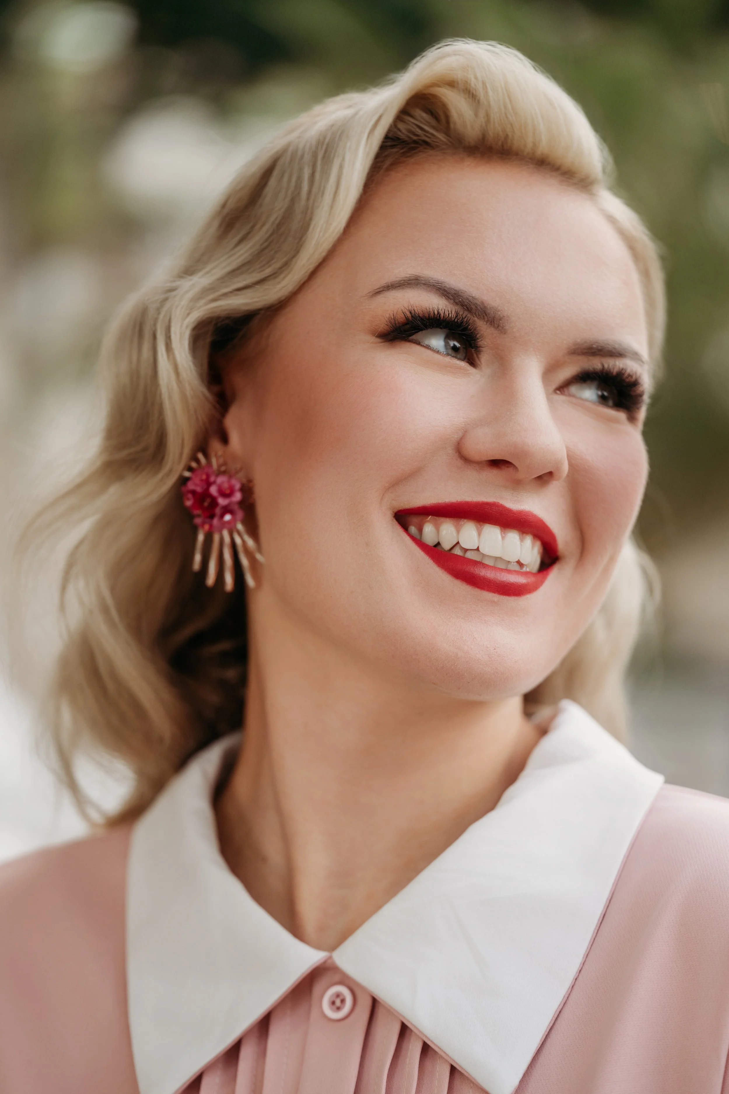 Nahaufnahme eines lachenden Frauenkopfes mit blonden, gewellten Haaren, rotem Lippenstift und pinken Ohrringen, draußen.