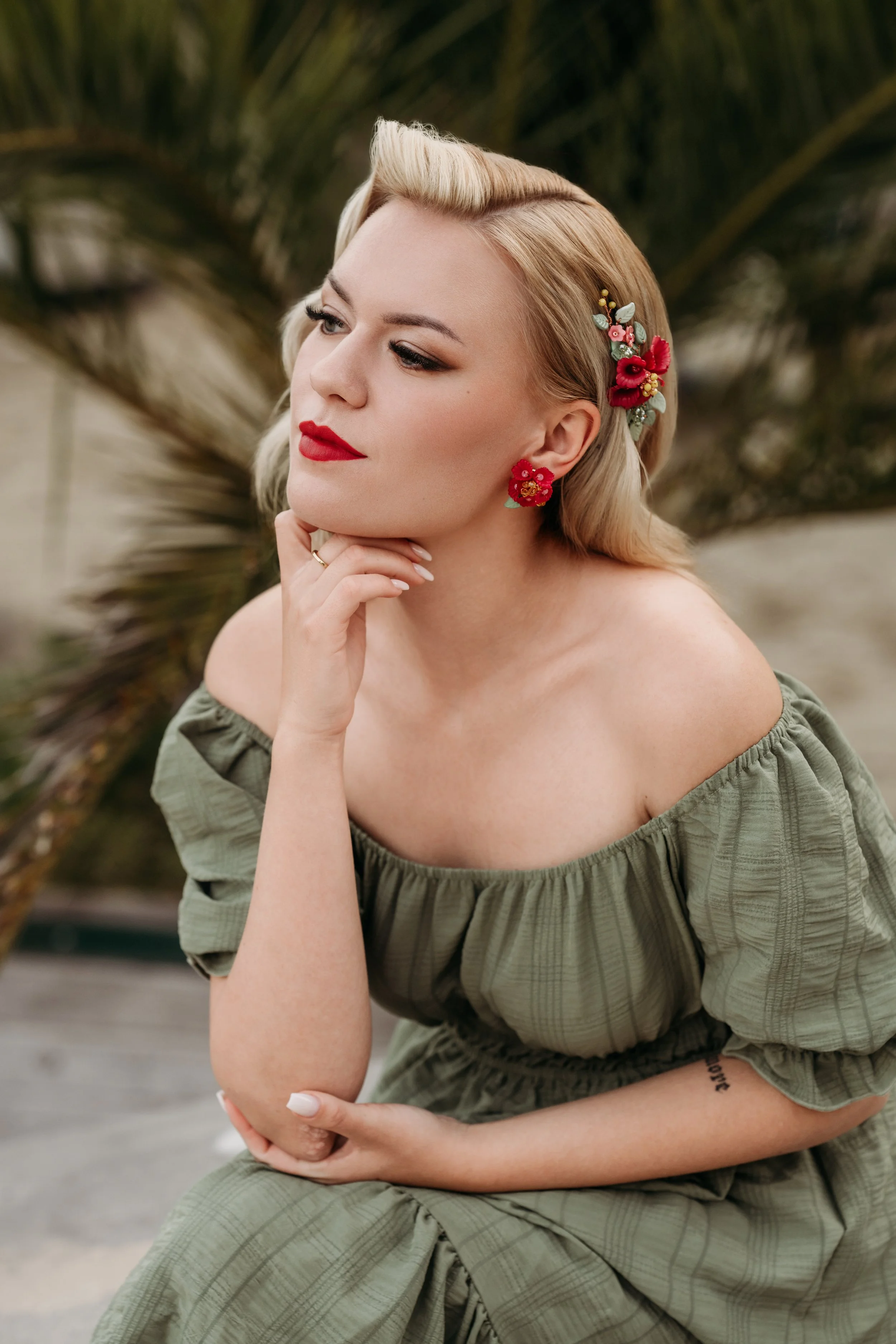 Eine Frau mit blondem Haarschopf, rotem Lippenstift und floralen Ohrringen, trägt ein grünes, off-shoulder Kleid, sitzt vor einer Pflanze.