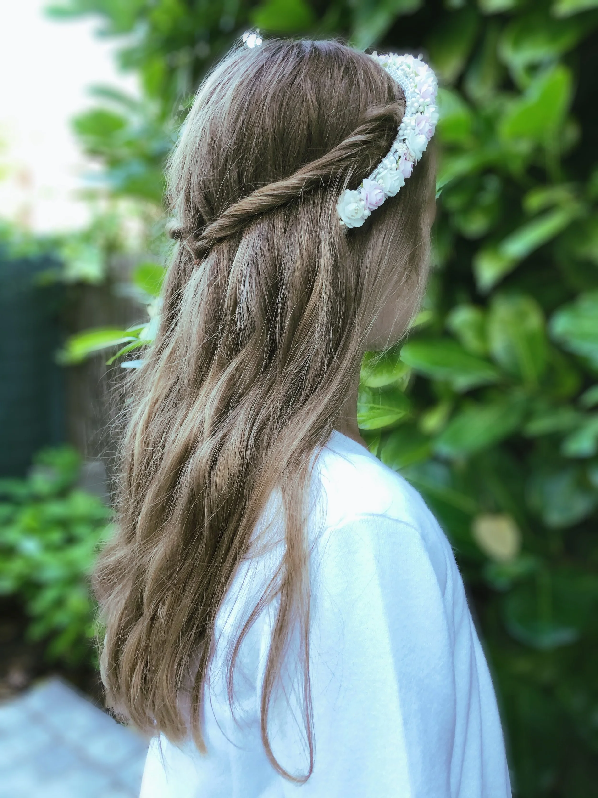 Ein Mädchen mit braunen, lockigen Haaren trägt eine Blütenkrone und einen weißen Pullover, steht vor grünen Blättern im Hintergrund.