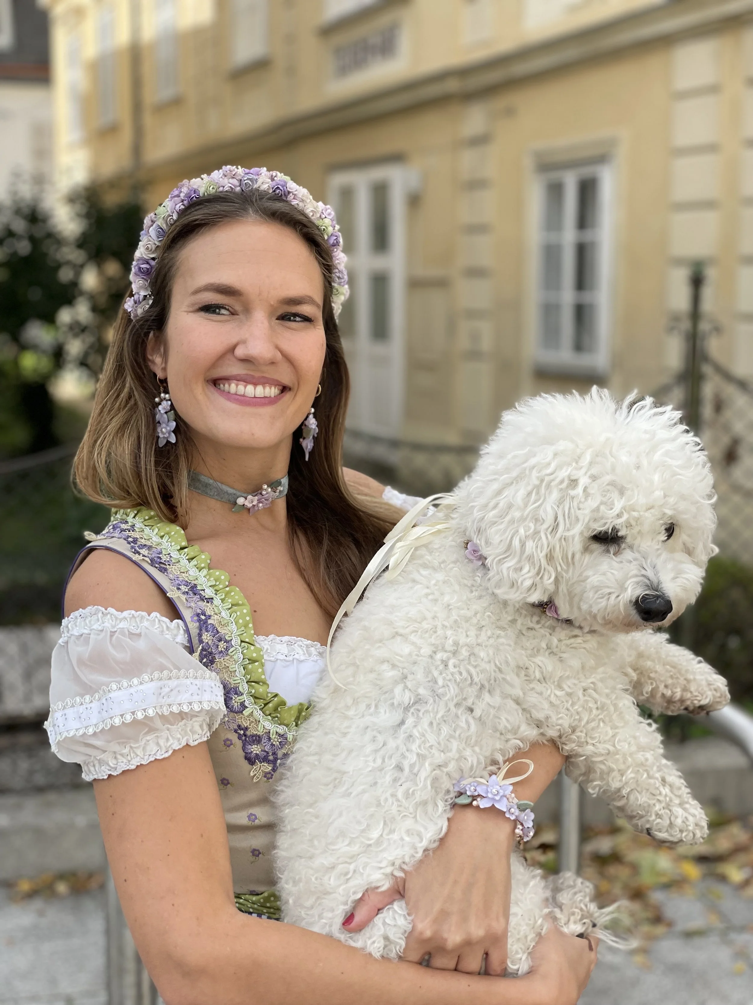 Eine lachende Frau in traditionellem Kleid hält einen weißen, lockigen Hund in den Armen, draußen vor einem Haus.