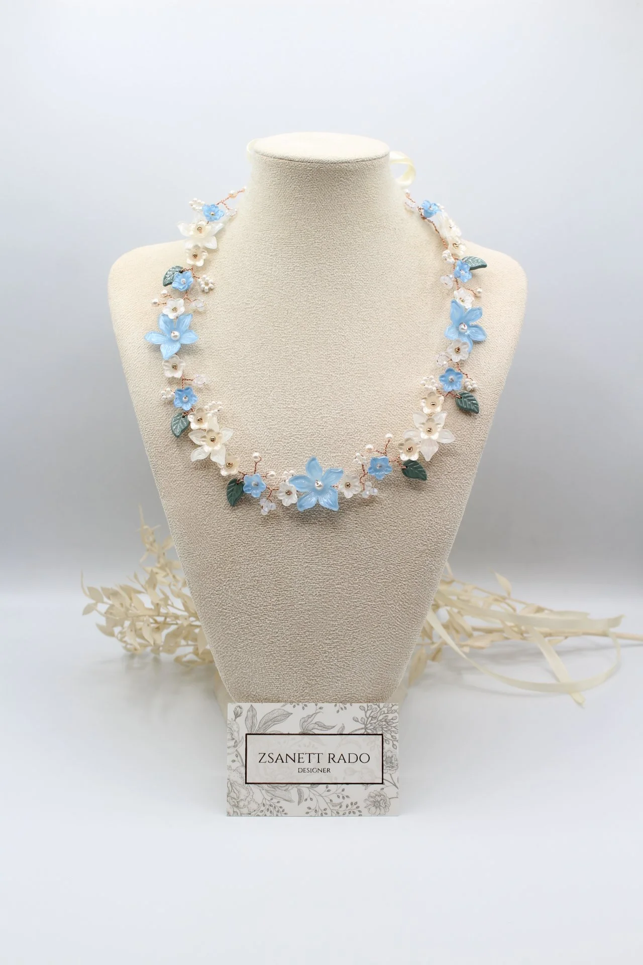 Halskette mit blauen und weißen Blumen, auf einem Schmuckdisplay mit Namensschild 'Zsanett Rado Designer'