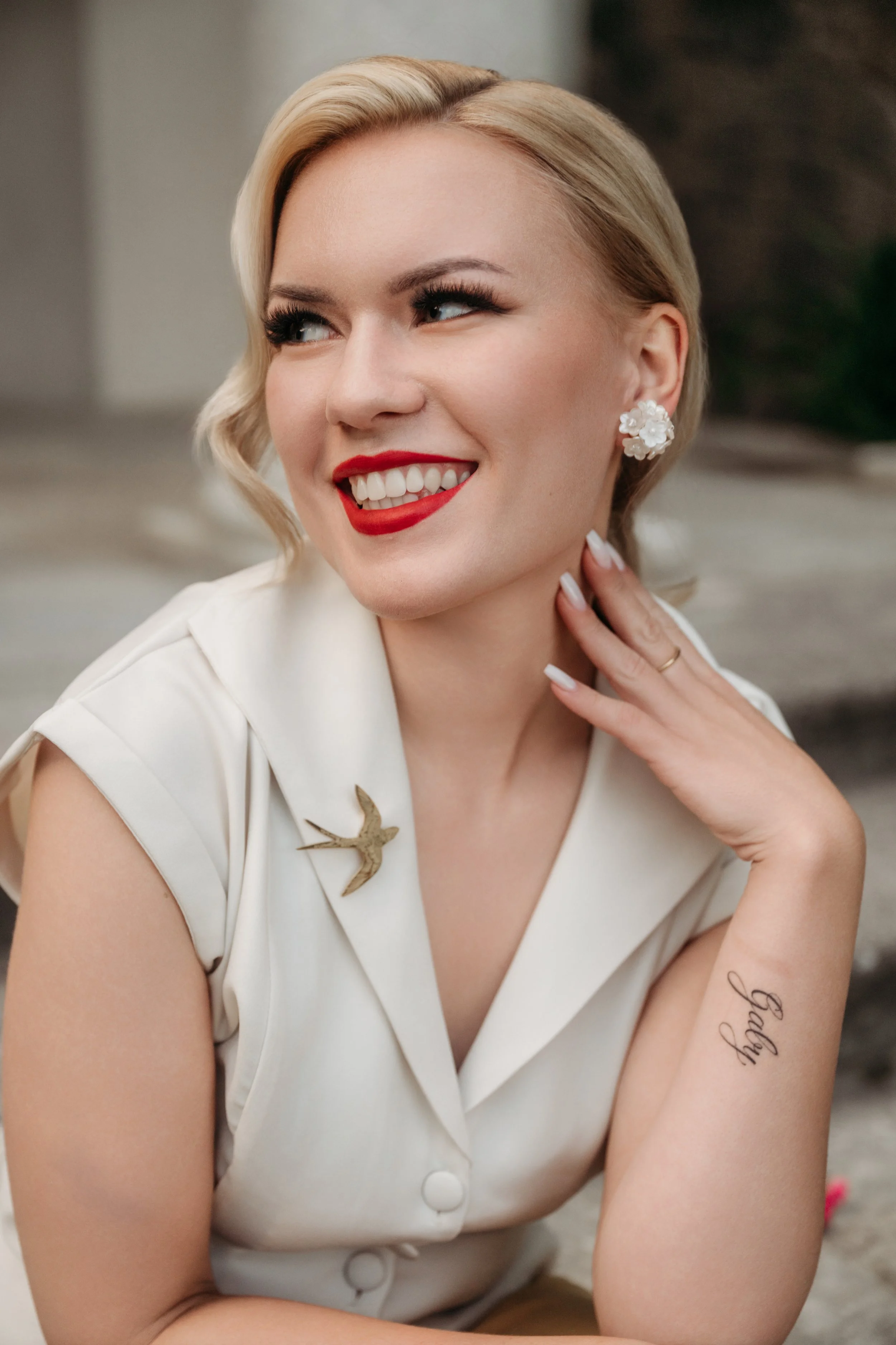 Eine lachende Frau mit blonden, welligen Haaren, rotem Lippenstift, weißen Zähnen und Ohrringen, trägt ein weißes Kleid mit einer Brosche in Form eines fliegenden Vogels und hat auf der Innenseite ihres rechten Unterarms die Tätowierung "Jelly".