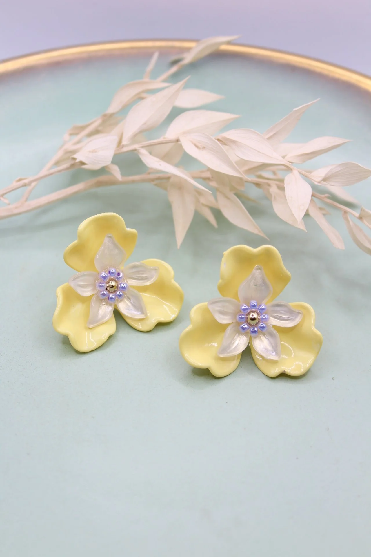 Gelbe Blumenohrringe mit klaren Blütenblättern und kleinen blauen Perlen in der Mitte, auf einem hellblauen Untergrund mit getrockneten weißen Zweigen.