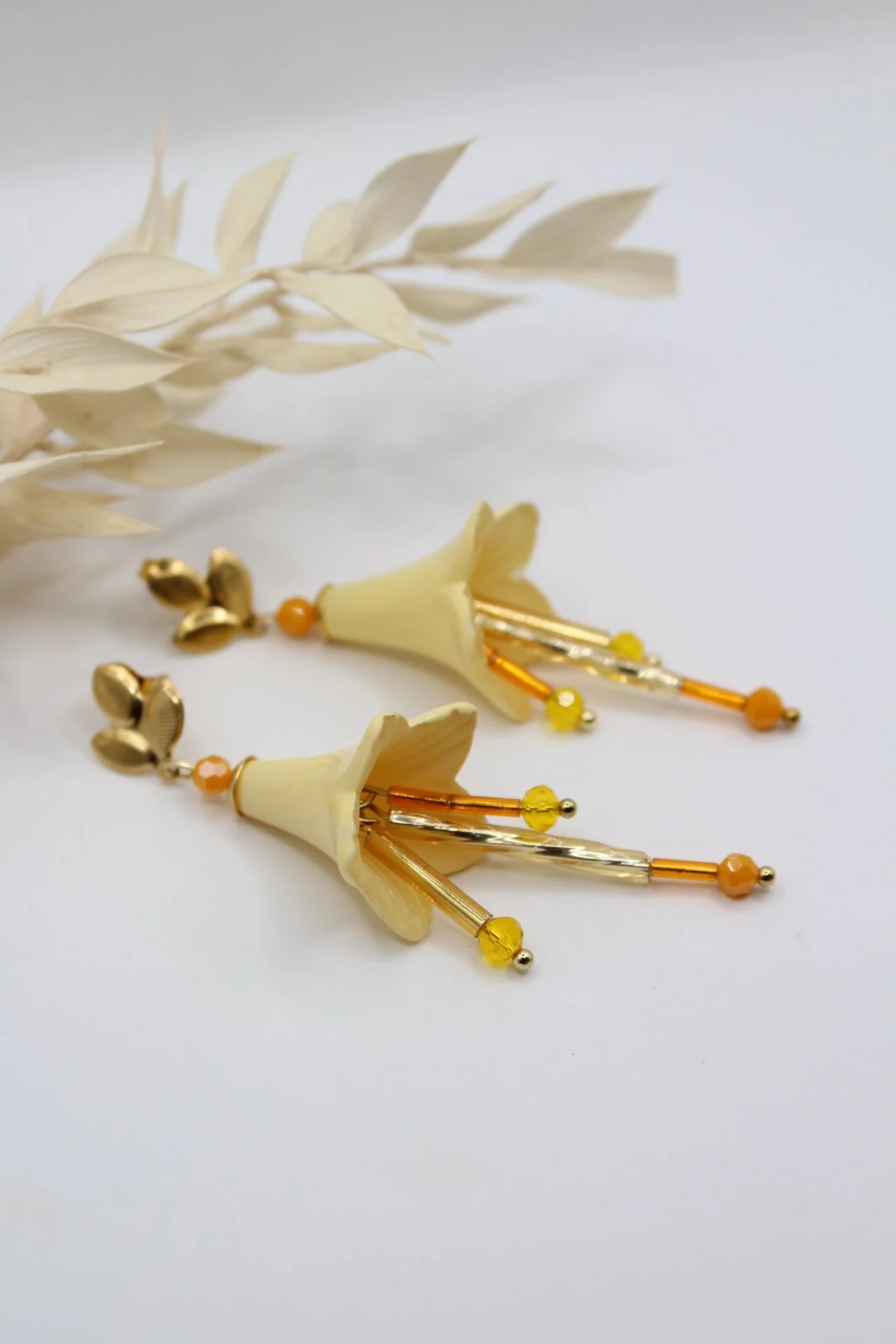Ein Paar Ohrringe mit gelben Blumenmotiven, goldfarbenen und orangen Akzenten, auf weißem Hintergrund neben getrockneten weißen Blättern.