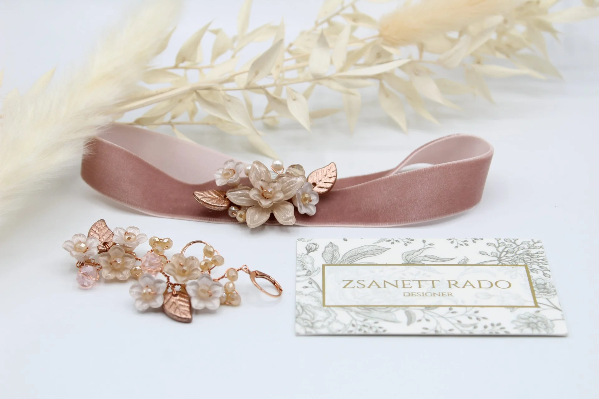 Schmuckset aus rosa und weißen Blumen, Ohrringe und eine Brosche, ein rosa Samthalsband mit Blumen Dekoration, ein Visitenkarten mit Schriftzug 'Zsanett Rado Designer', weiße dekorative Pflanzen im Hintergrund.