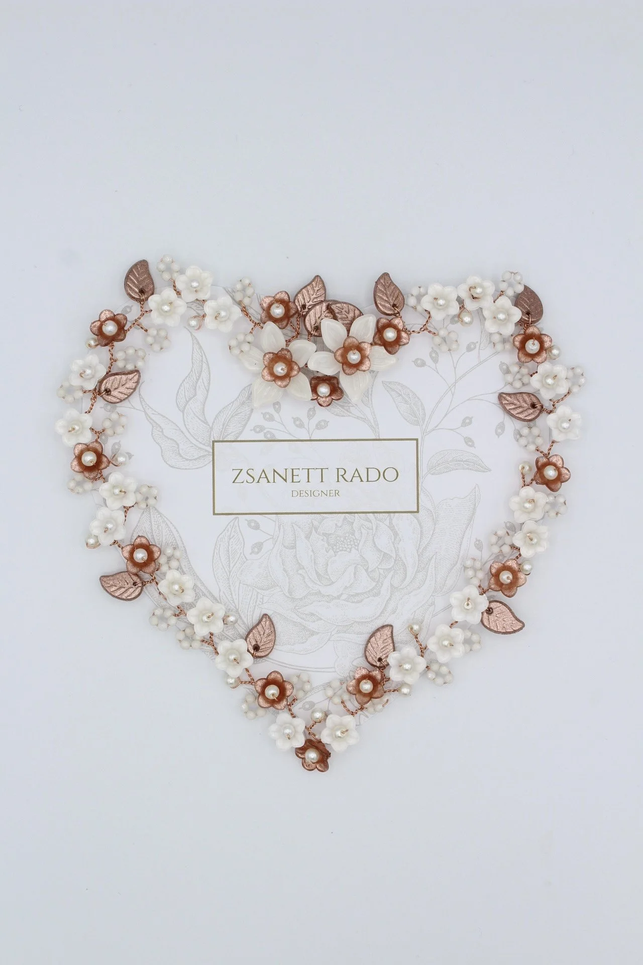 Ein Herz aus kleinen weißen und roséfarbenen Blumen und Blättern, mit einem Kärtchen in der Mitte, darauf steht 'Zsanett Rado Designer'.