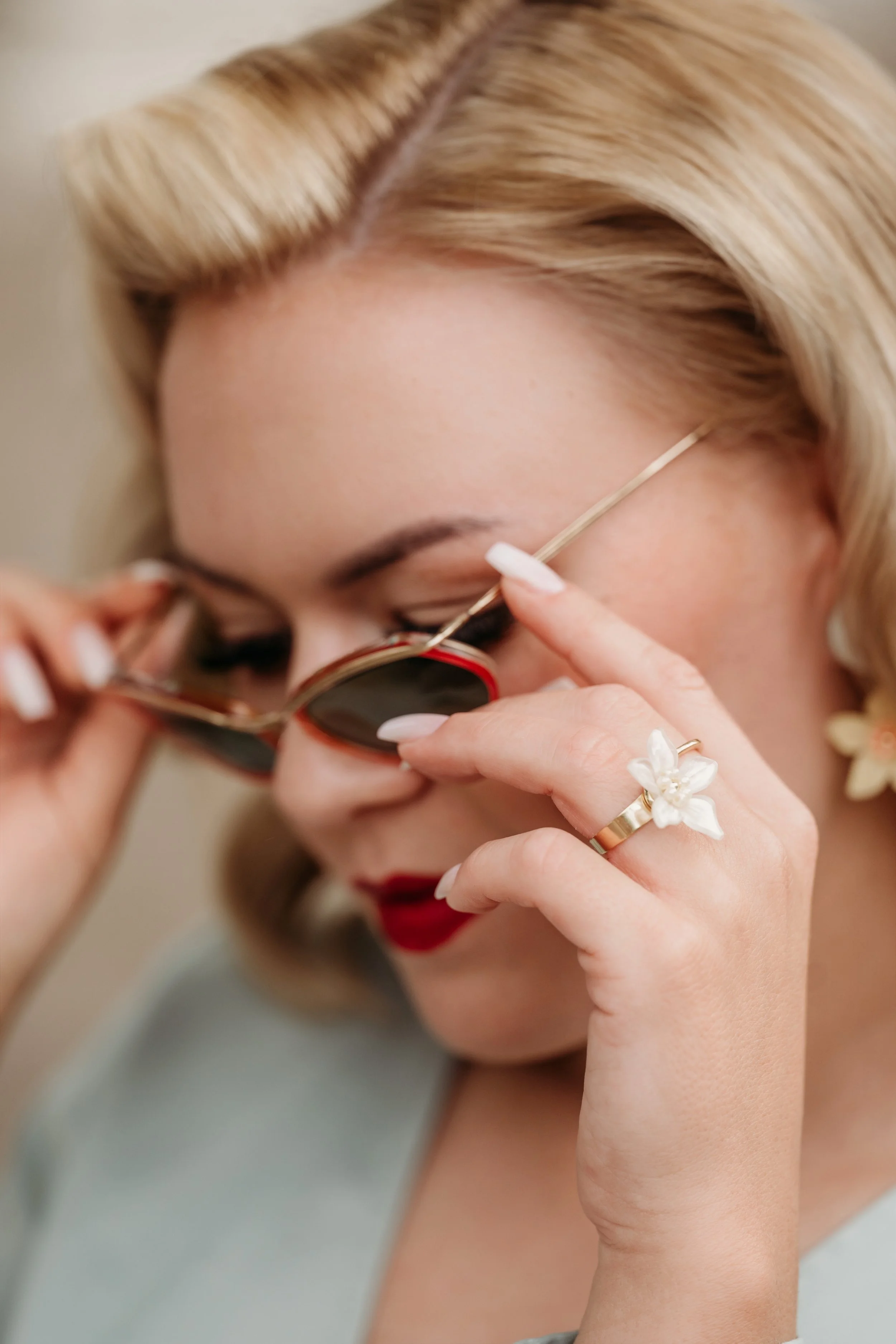Frau mit blondem Haar, rote Lippen, Sonnenbrille und Ohrringen, die eine Brille absetzt.
