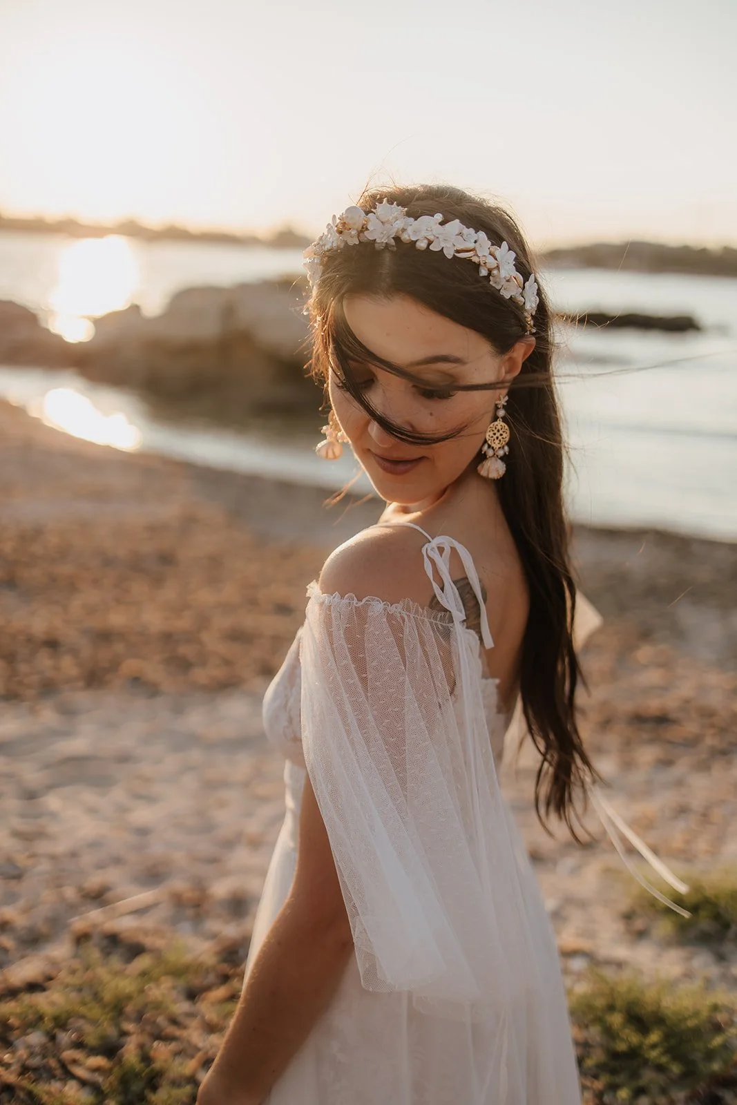 Junge Frau in weißem Sommerkleid mit Blumenkranz am Strand bei Sonnenuntergang.