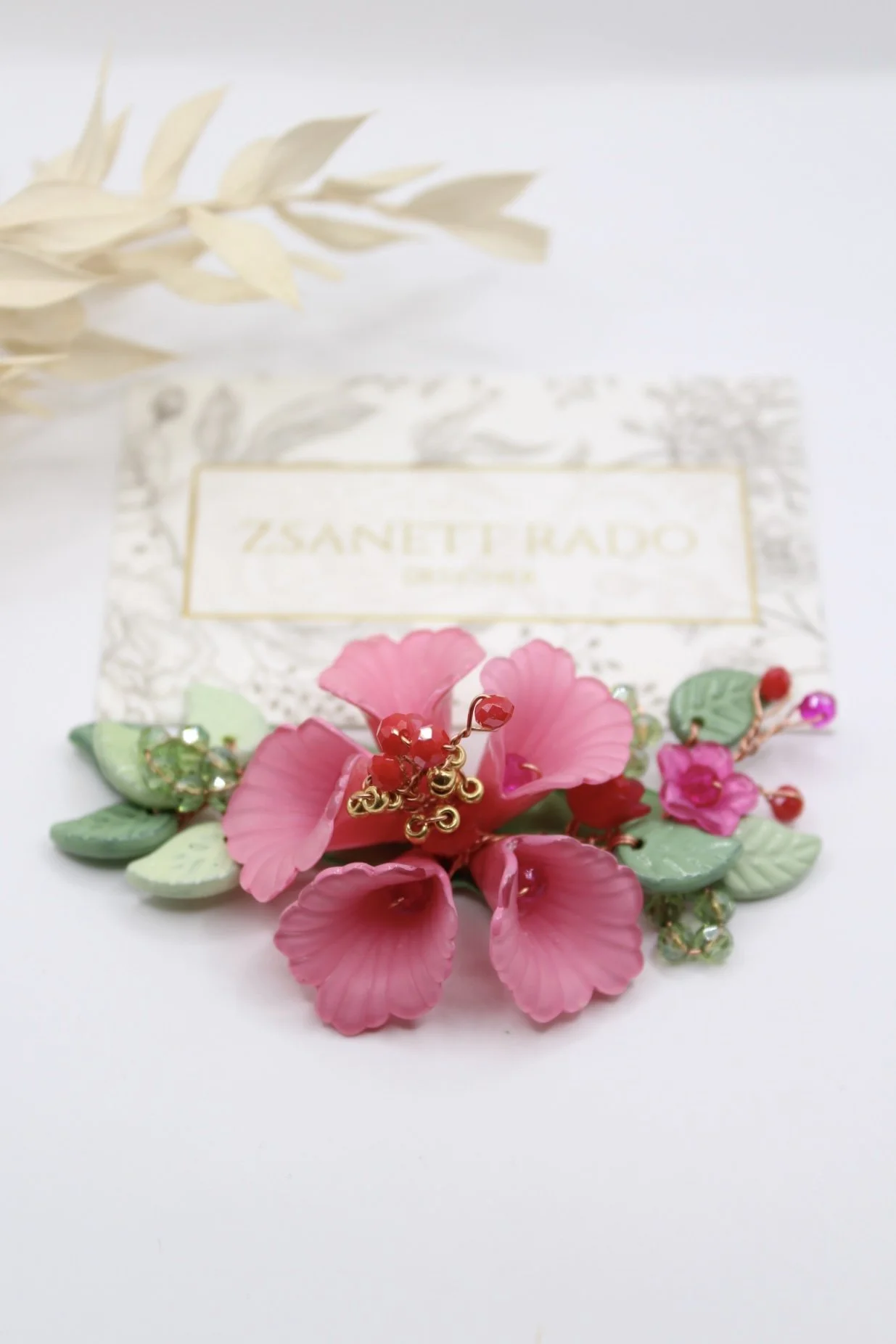 Halskette mit rosa Blüten und roten Beeren, liegend auf einem weißen Hintergrund, mit Verpackung im Hintergrund.