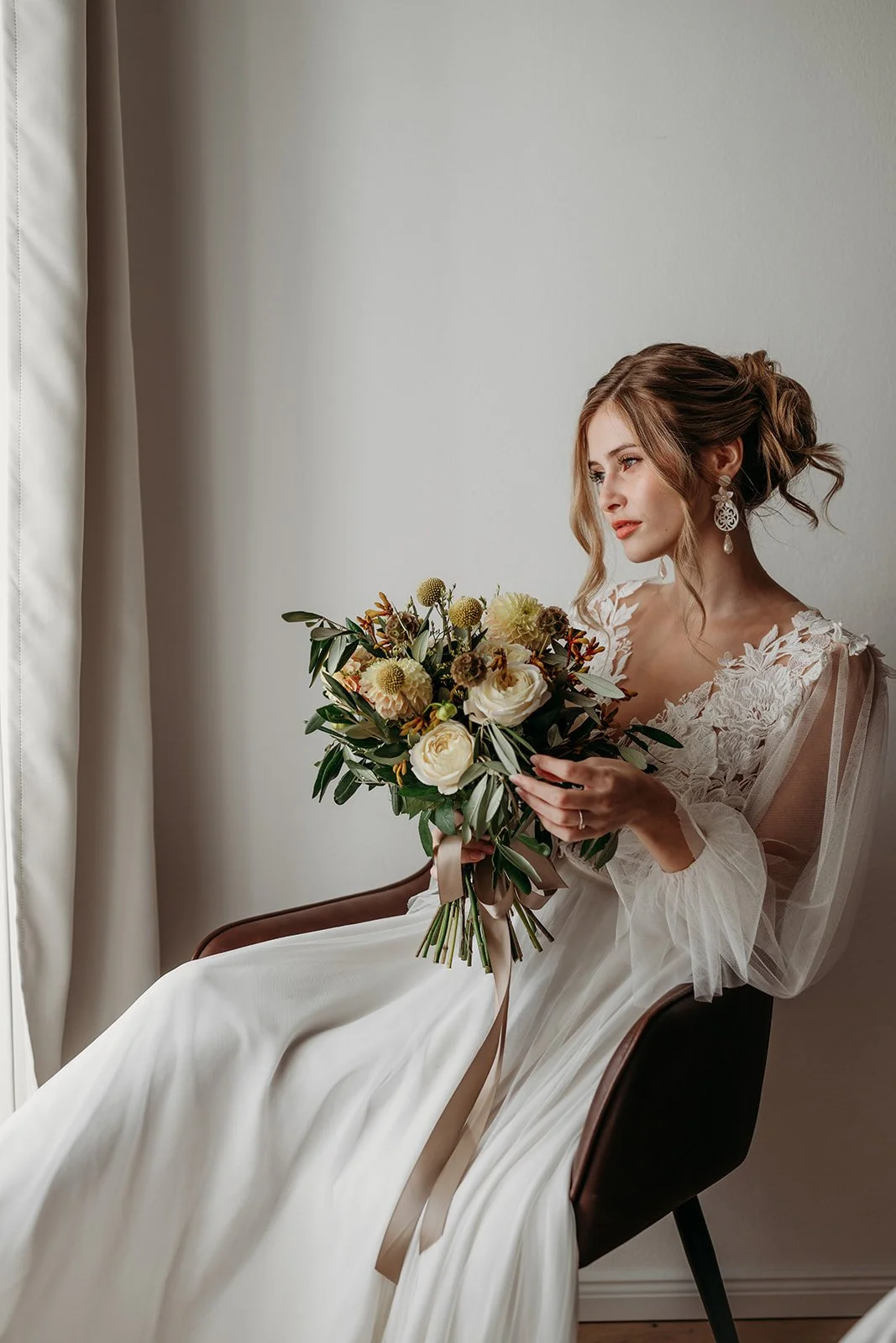 Eine junge Frau in einem weißen Brautkleid sitzt an einem Fenster und hält einen Brautstrauß mit weißen und grünen Blumen.