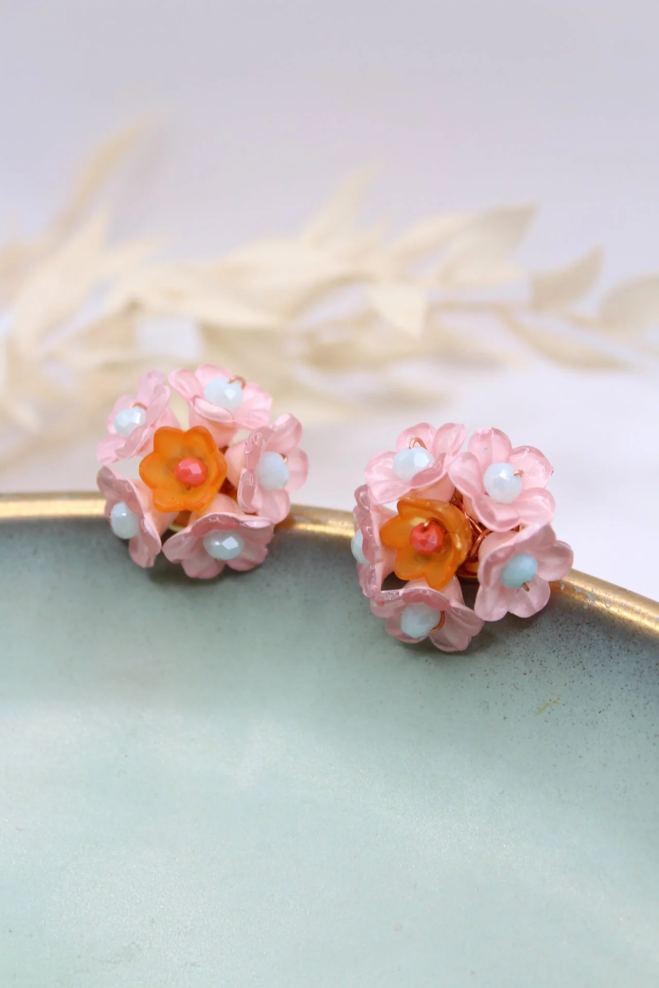 Zwei Ohrringe in Form von blühenden Blumen mit pinken, weißen und orangefarbenen Details auf einer hellgrünen Oberfläche, mit weißem dekorativem Zweig im Hintergrund.