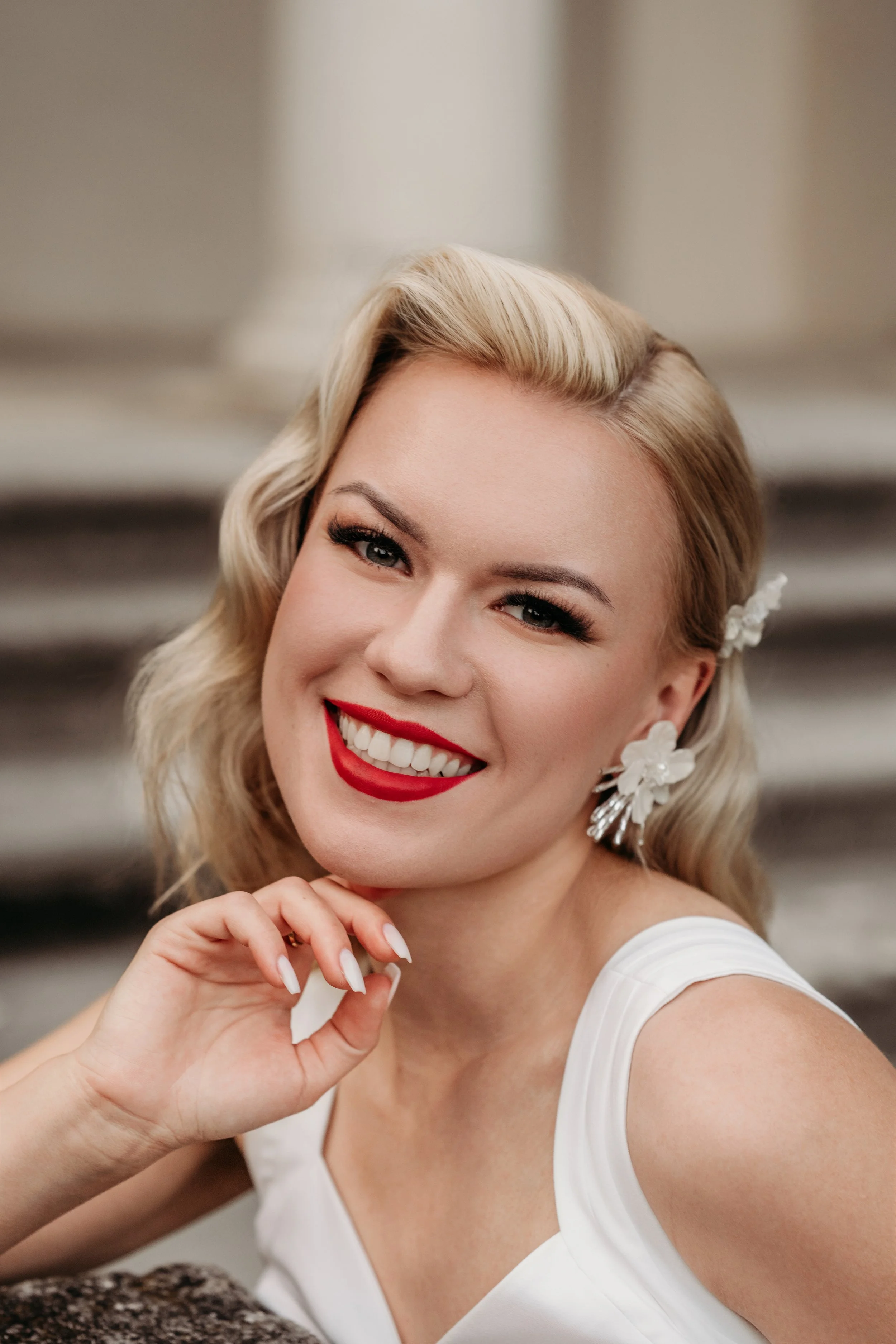 Eine lachende blonde Frau mit rotem Lippenstift und weißen Zähnen, trägt weiße Kleidung und große weiße Blumenohrringe, posiert vor einer neutralen Wand.