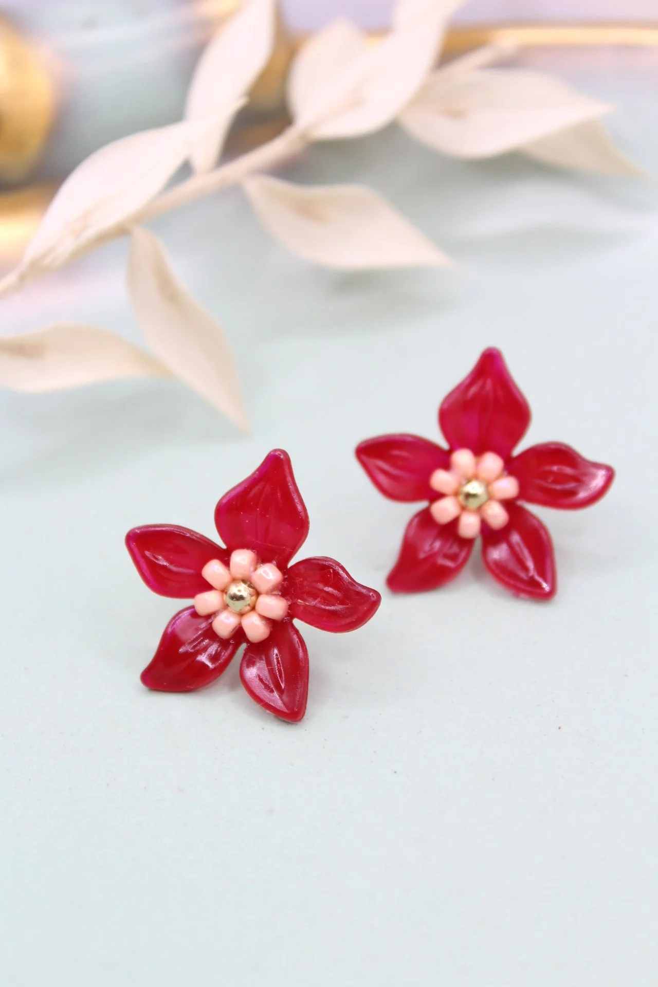Rote Blumenohrringe mit pinken Akzenten und kleinen weißen Blüten in der Mitte, auf hellblitem Hintergrund mit weißen Blättern im Hintergrund.